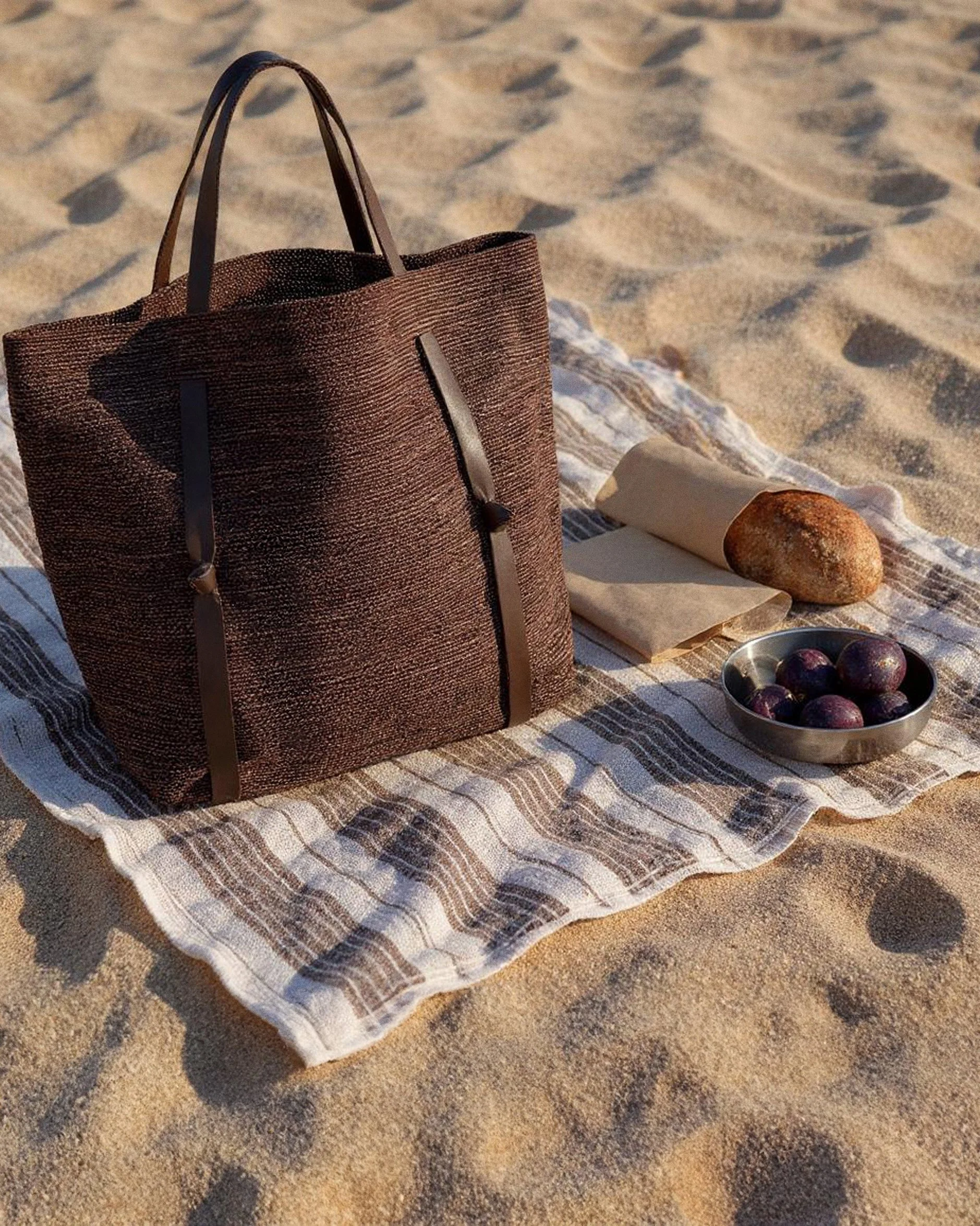 A brown tote bag resting on a striped beach towel on sandy ground, alongside a white rolled-up cloth, a round loaf of bread, and a small metal bowl filled with purple grapes.