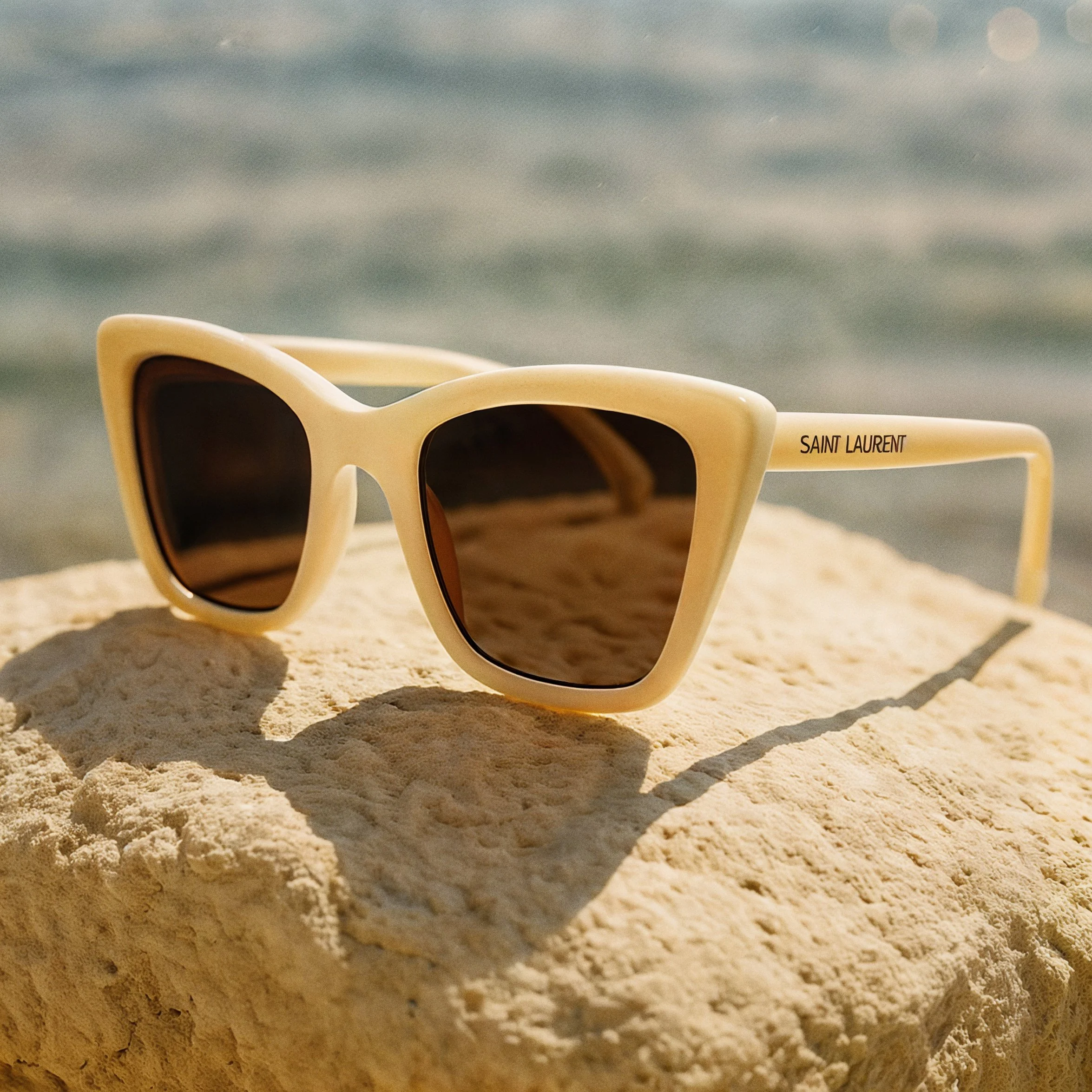 Close-up of a pair of Saint Laurent sunglasses with beige frames, placed on sandy ground with a blurred background and sunlight.