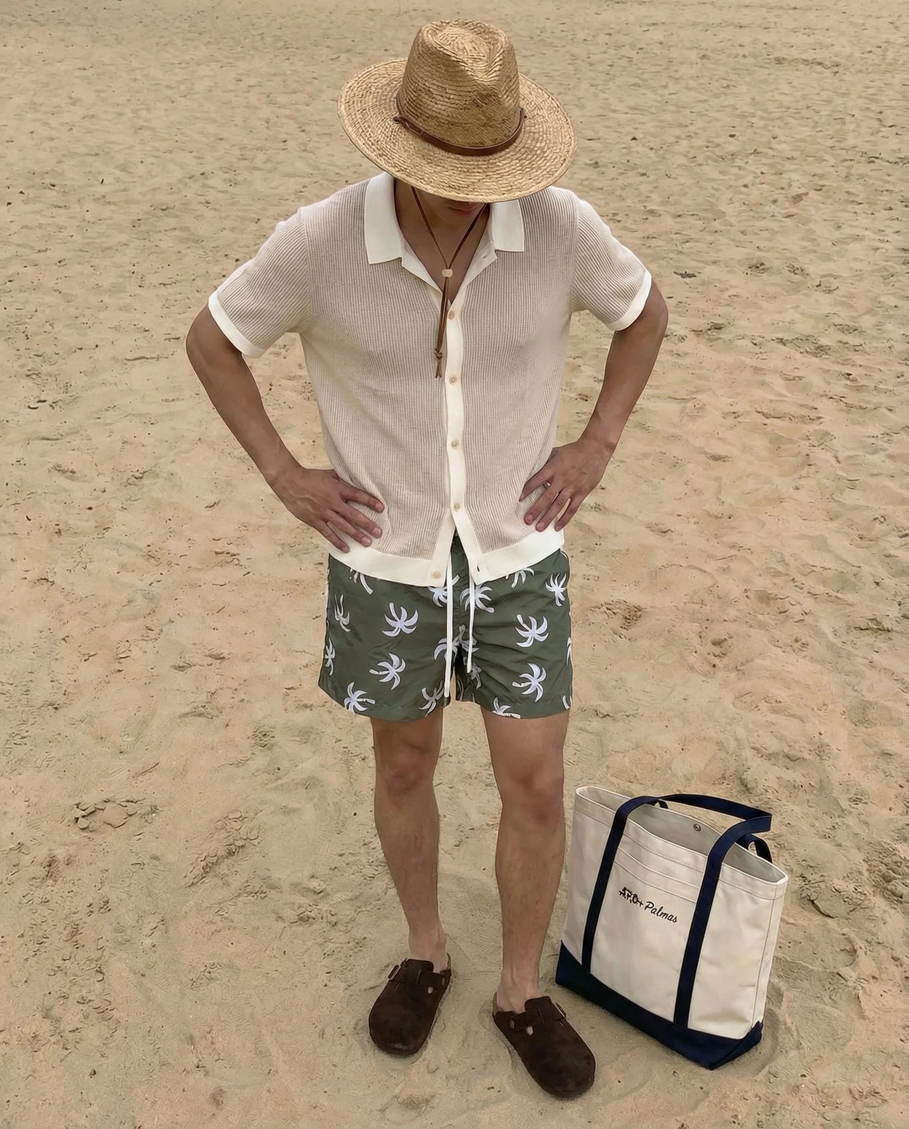 A person standing on a sandy beach, wearing a wide-brimmed straw hat, a beige short-sleeved shirt, patterned swim shorts, and sandals. The person is holding their hands on their hips, with a large white and navy tote bag on the sand nearby.