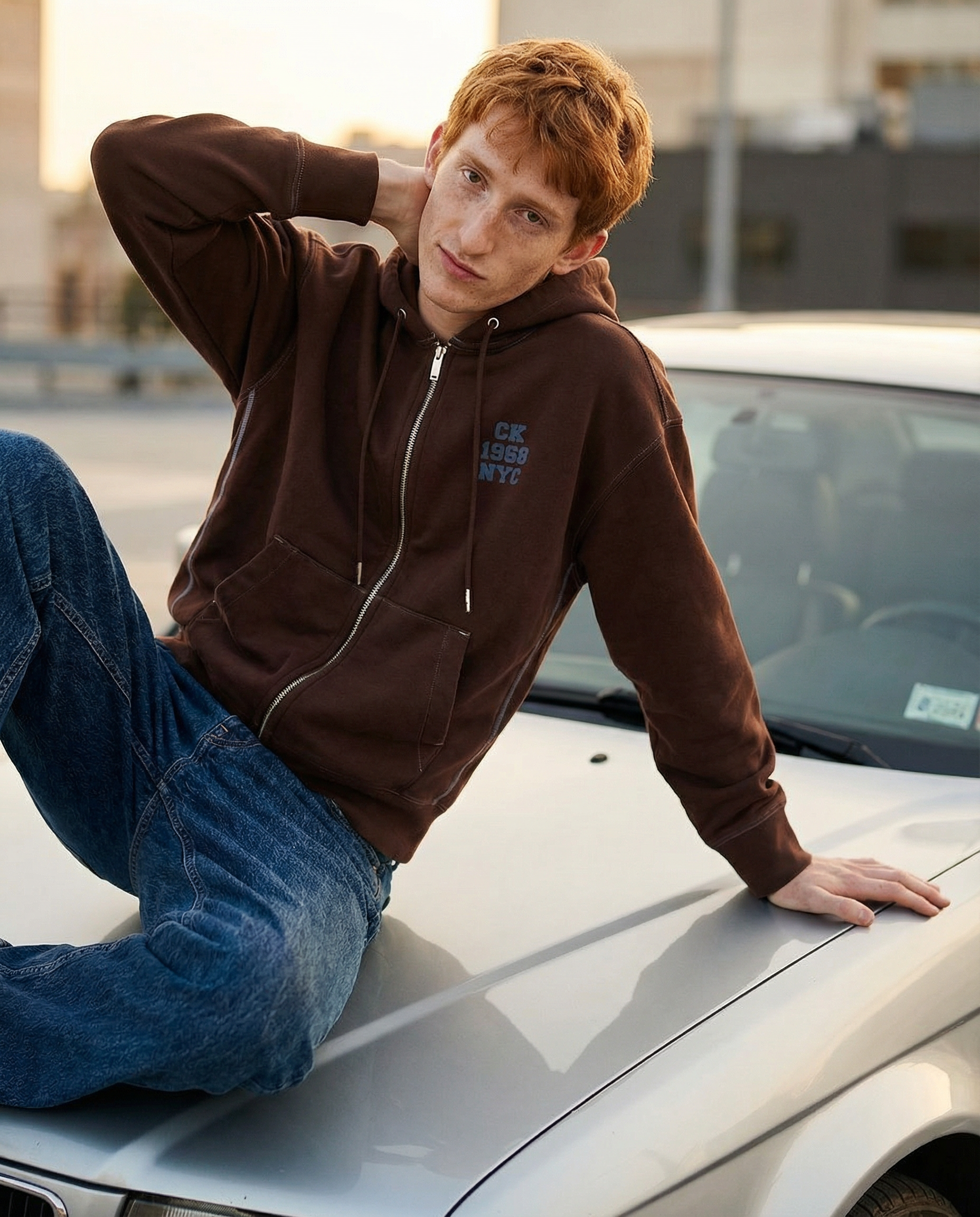 A young man with red hair and freckles, wearing a brown hoodie and blue jeans, leaning against a silver car with one hand on the hood and the other behind his neck, outdoors during daytime.