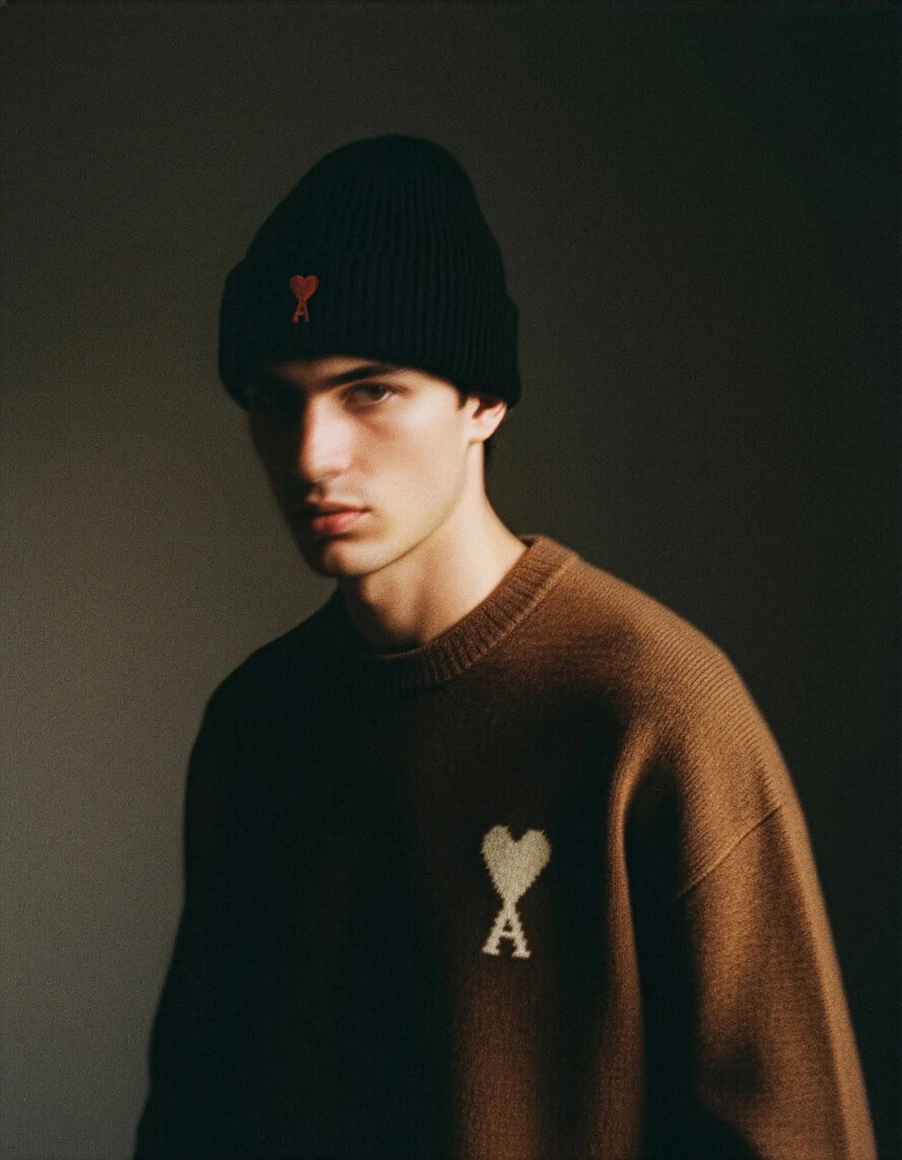 Close-up photo of a young man wearing a black beanie with a red heart and letter 'A' on it, and a brown sweater with a white heart and 'A' design on the front, against a plain dark background.
