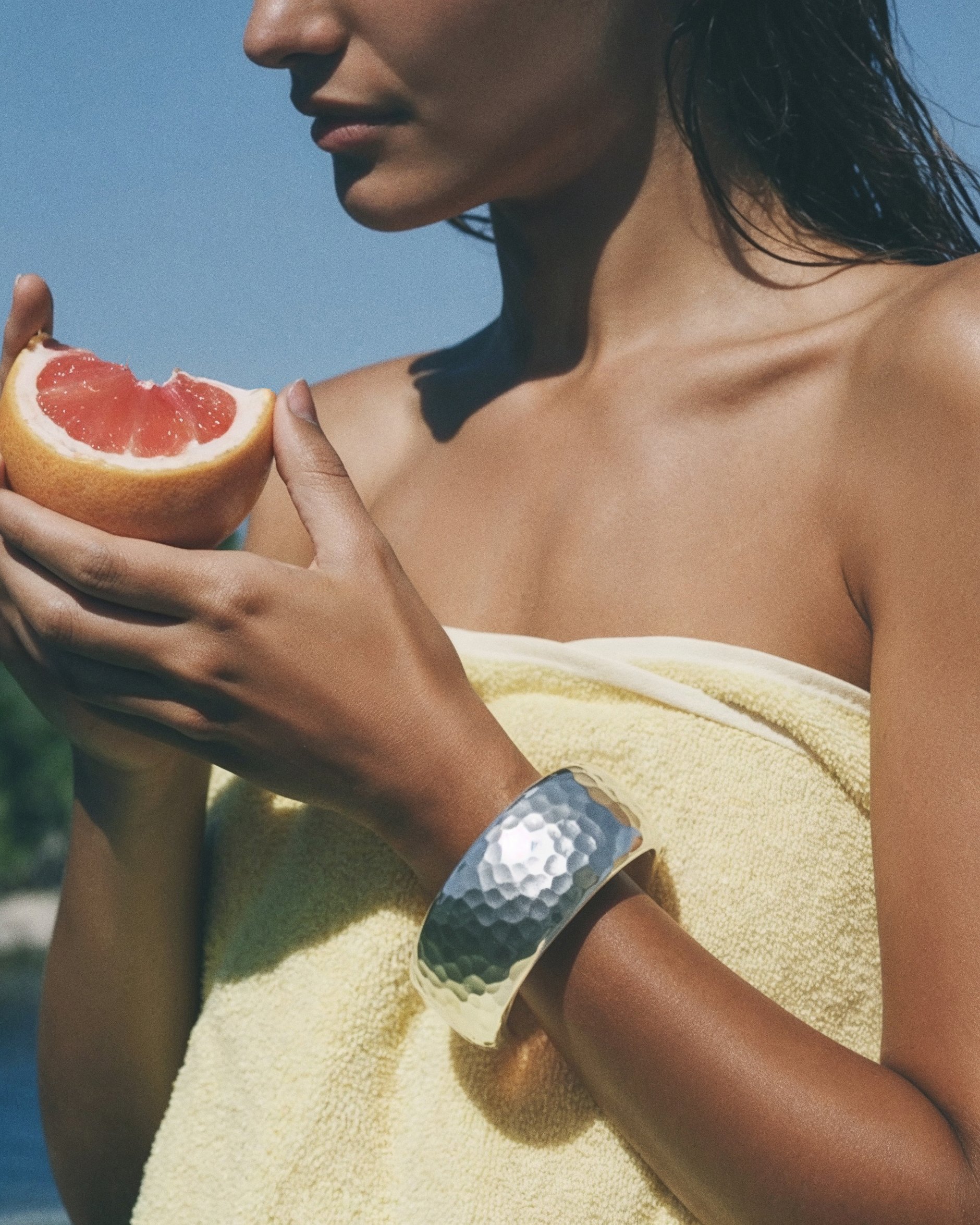 A woman with dark hair and tan skin holding a slice of grapefruit against a blue sky background, wrapped in a yellow towel, wearing a silver jewelry bracelet.