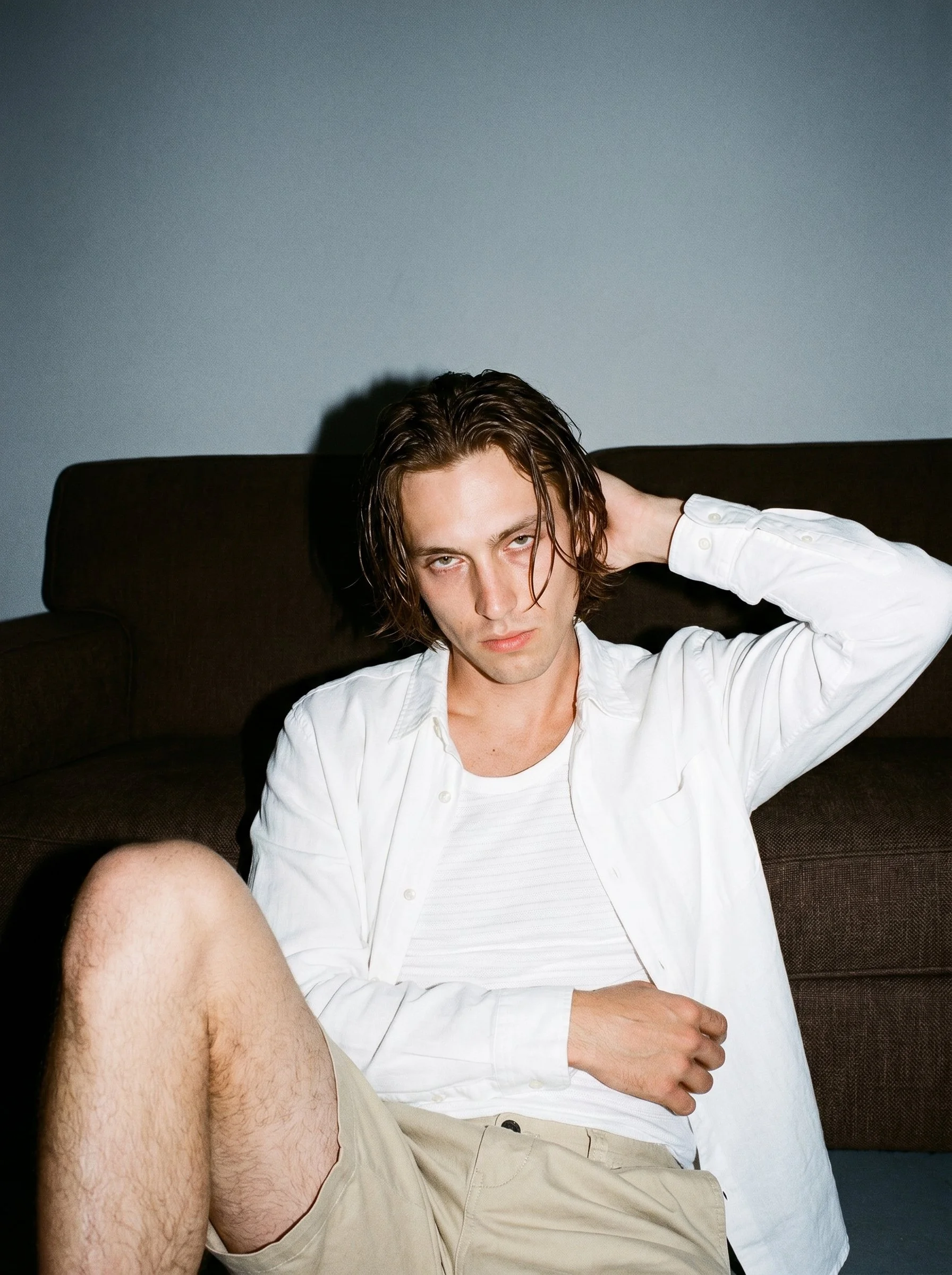 Young man with wet hair wearing a white shirt, sitting on a dark couch, with one leg up, in a dimly lit room with a plain background.