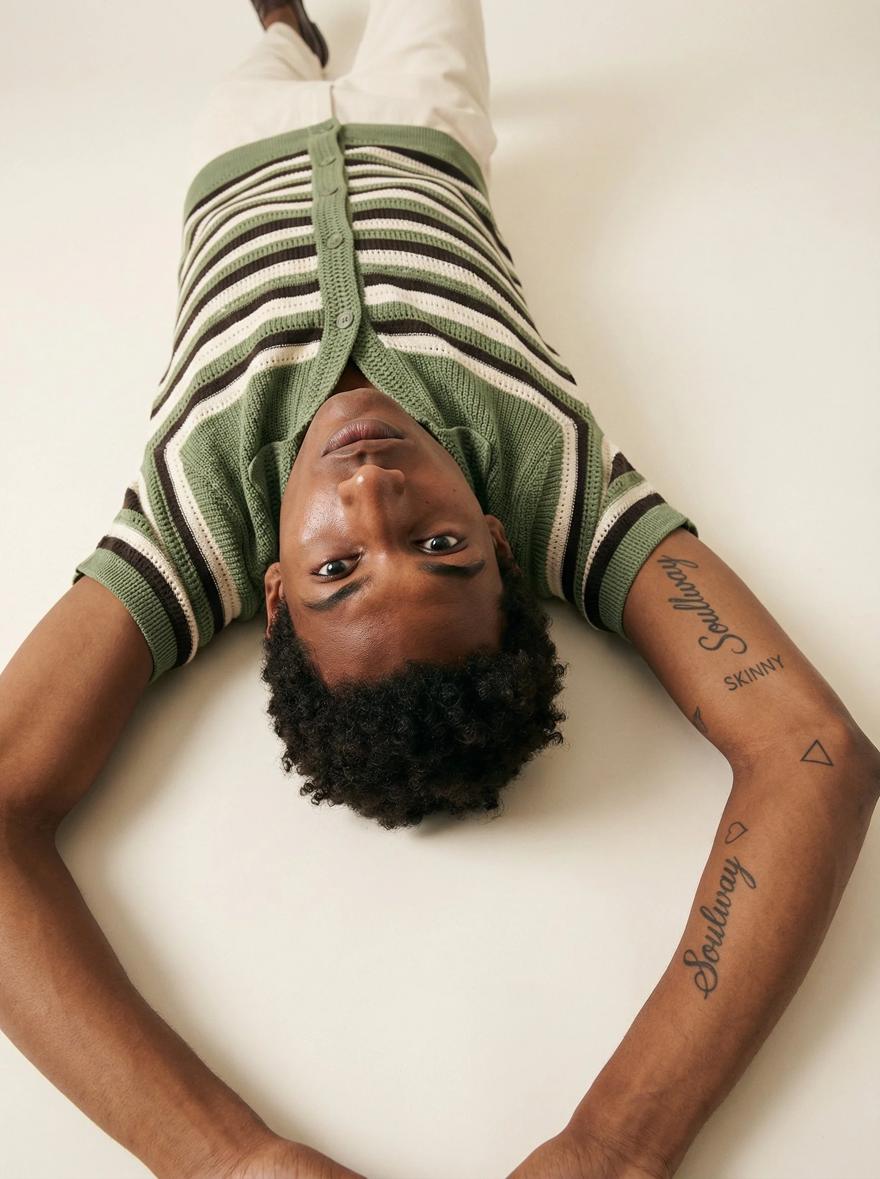 A person lying on a white surface, looking up at the camera. The person has dark curly hair, tattoos on their right arm, and is wearing a green, white, beige, and brown striped shirt.