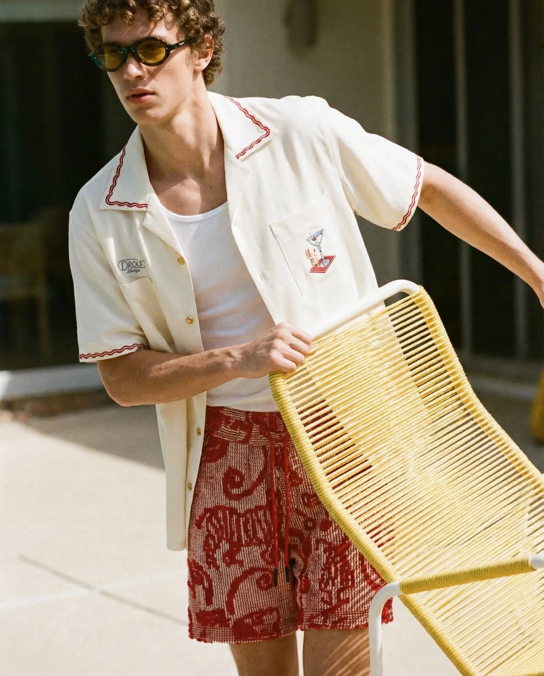 A young man with curly hair wearing yellow sunglasses, a white short-sleeve button-up shirt with red stitching, a white tank top underneath, and red patterned shorts, holding a yellow woven chair in an outdoor setting.