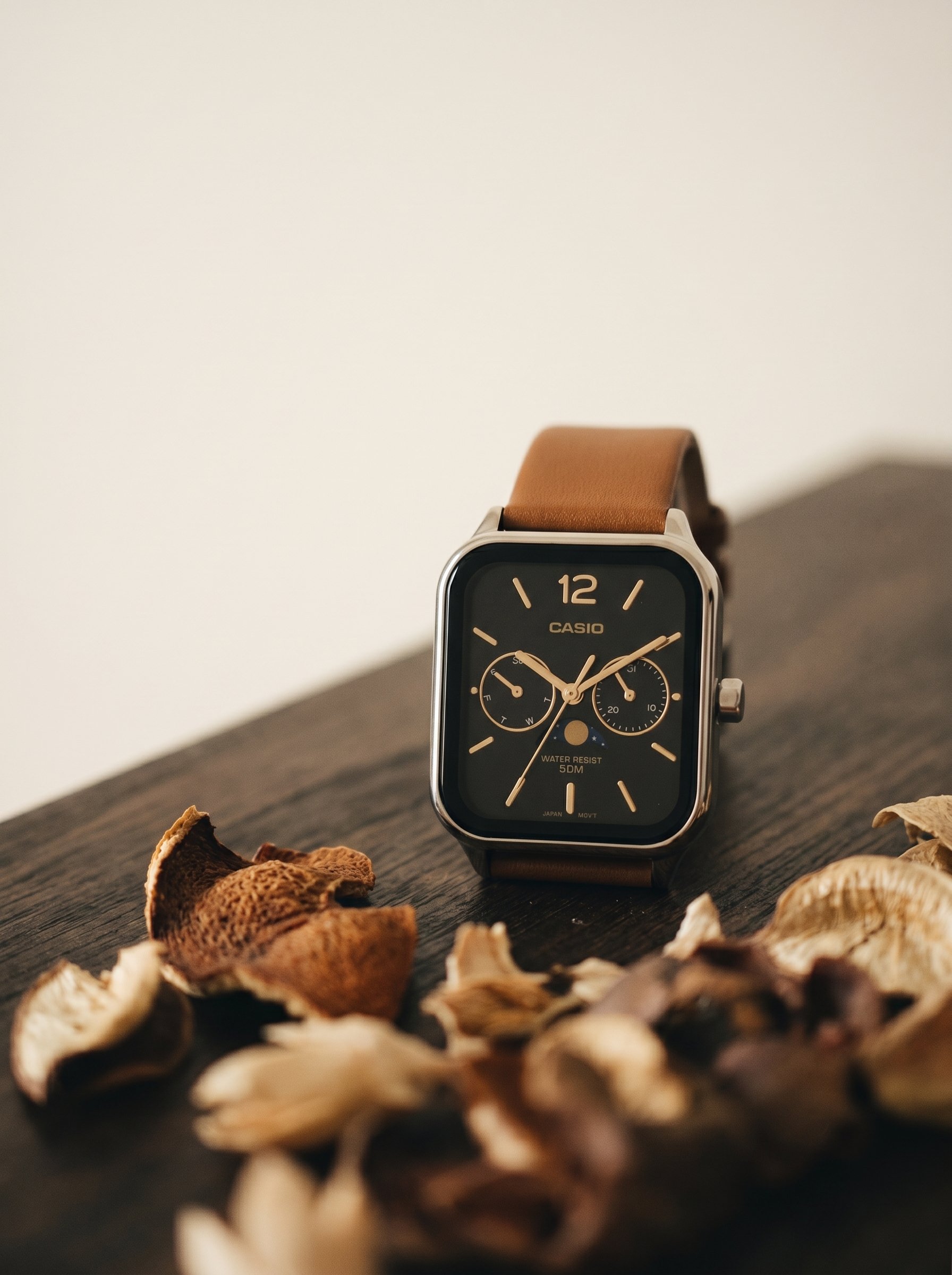 A smartwatch with a brown leather strap on a dark wooden surface surrounded by dried mushroom pieces.