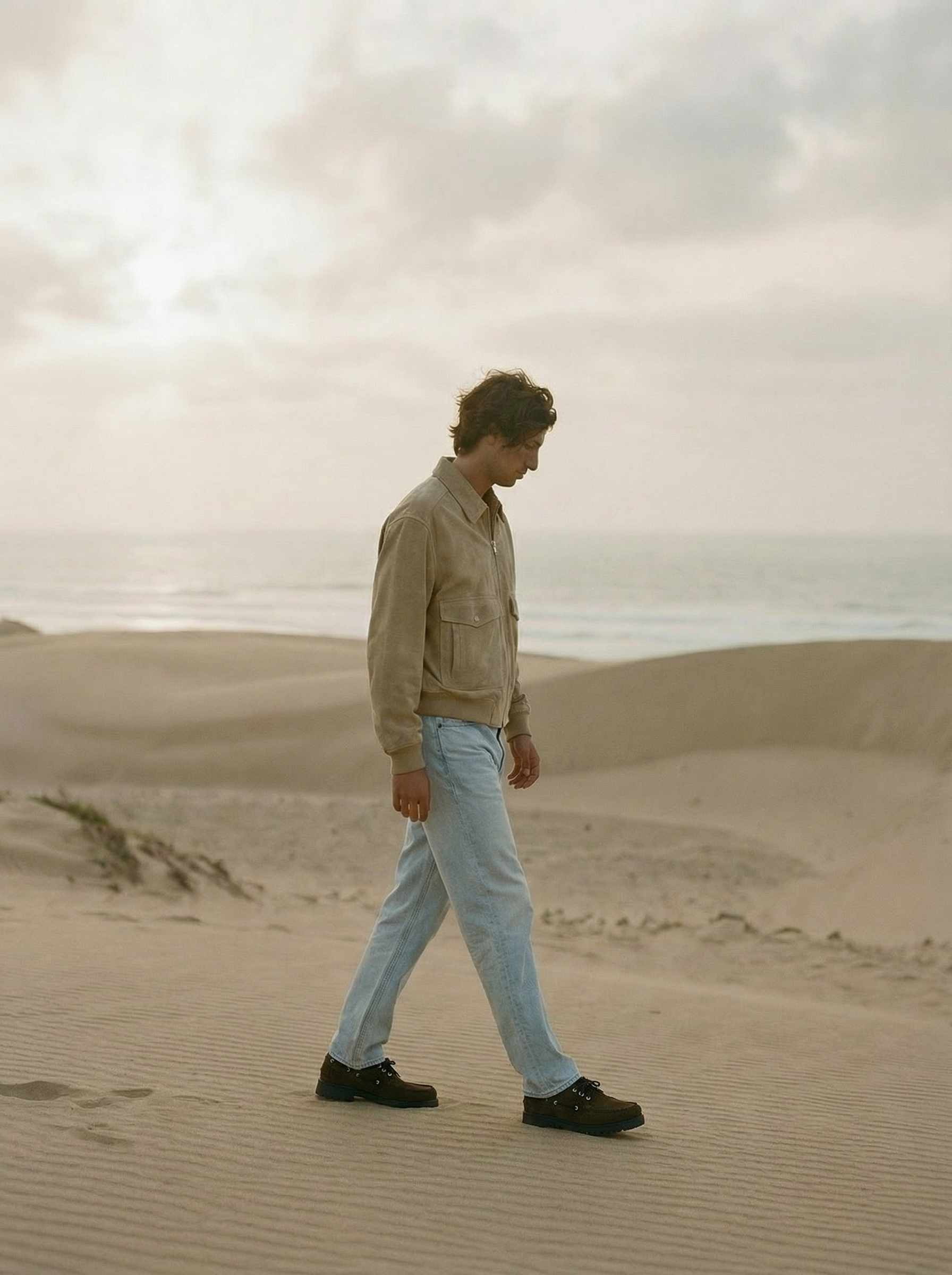 A young man with curly hair walking on a sandy beach with overcast sky in the background.  AI fashion editorial.
