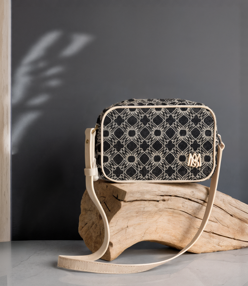 A patterned black and beige handbag with a beige strap, placed on a piece of driftwood against a dark gray wall.