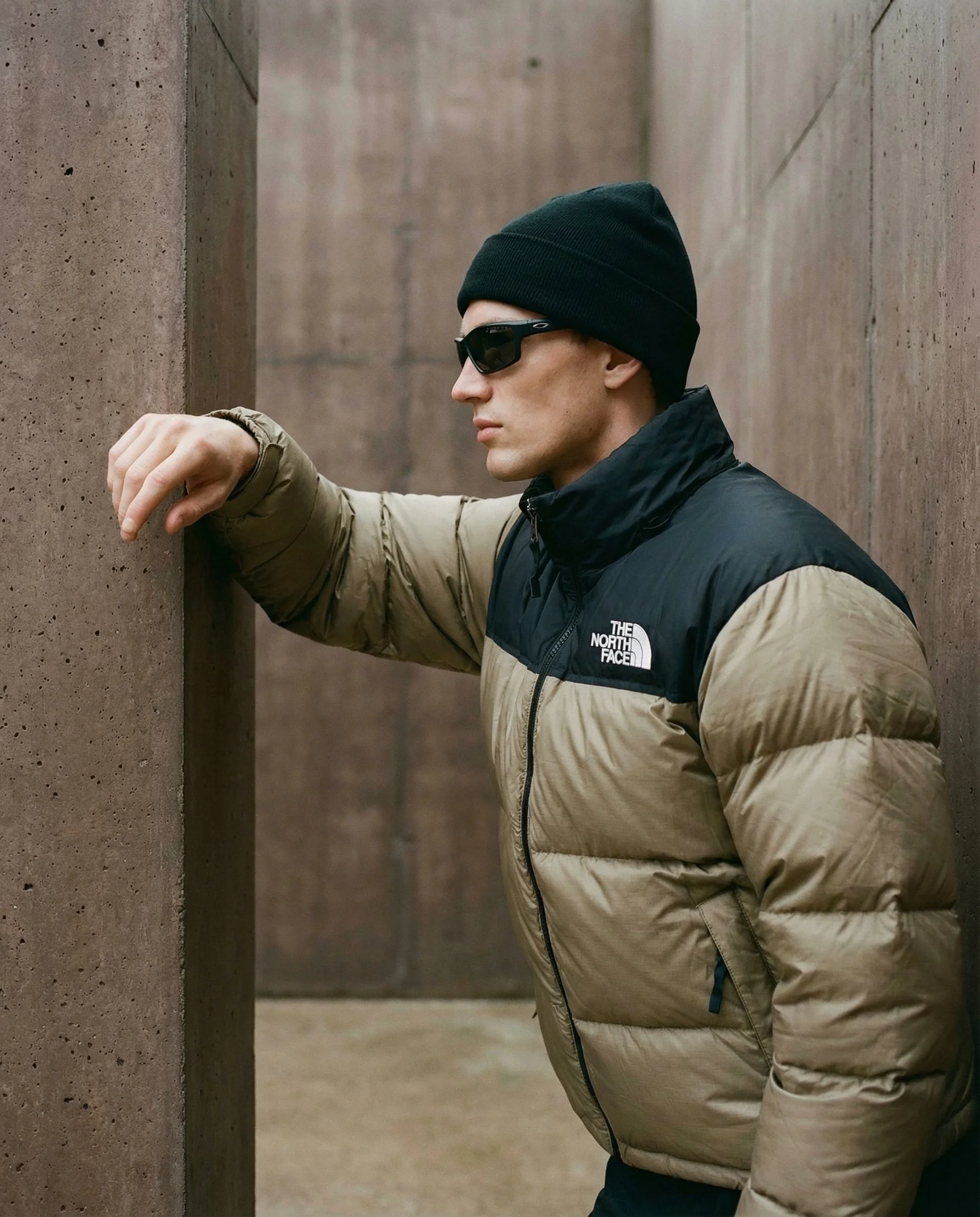 A man wearing sunglasses, a black beanie, and a two-tone puffer jacket leaning against a concrete wall with his arm raised and hand resting on the wall.