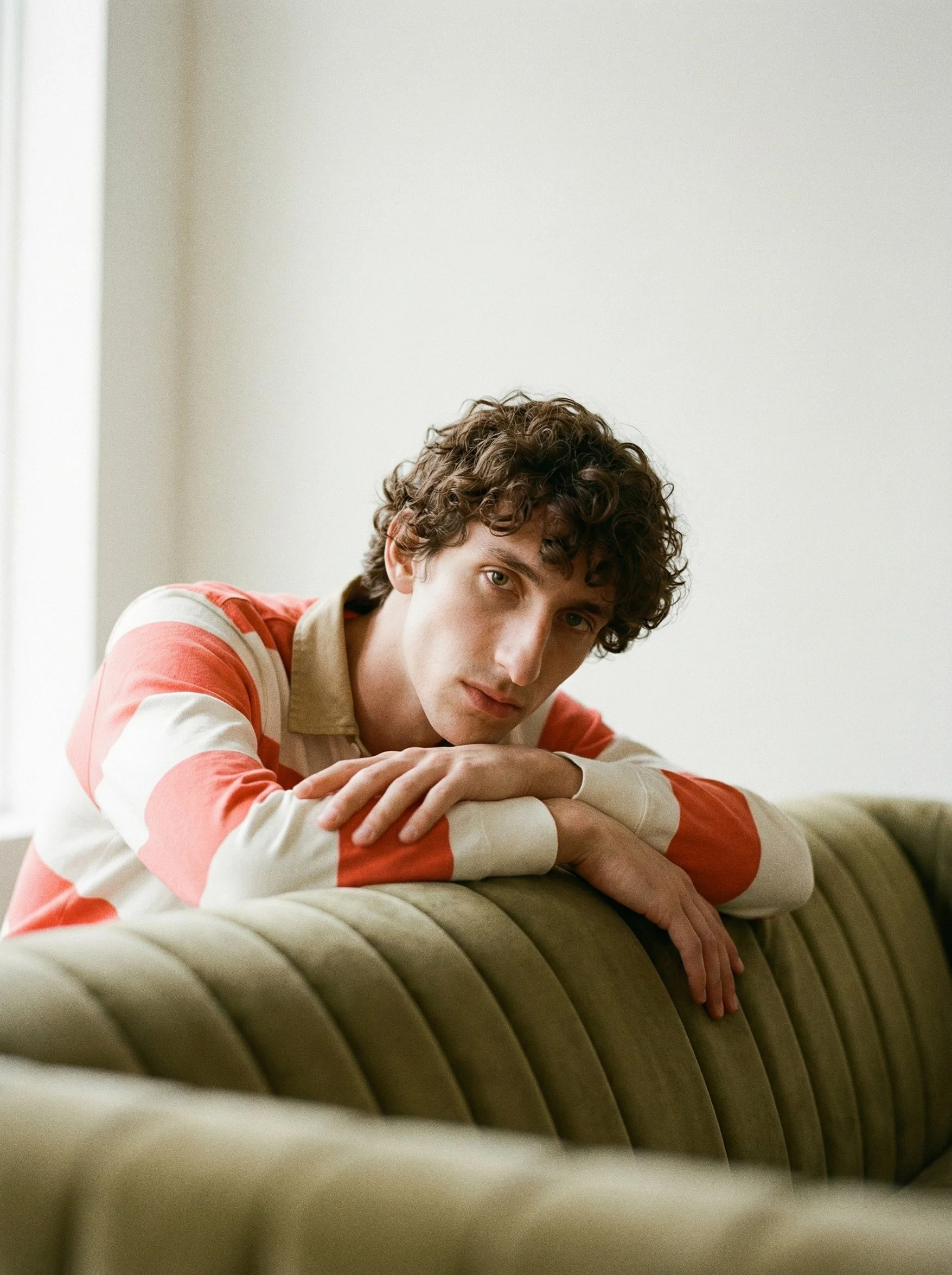 Young man with curly hair resting his head on his crossed arms on a beige couch, wearing a red and white striped shirt, in a room with a plain light-colored wall.