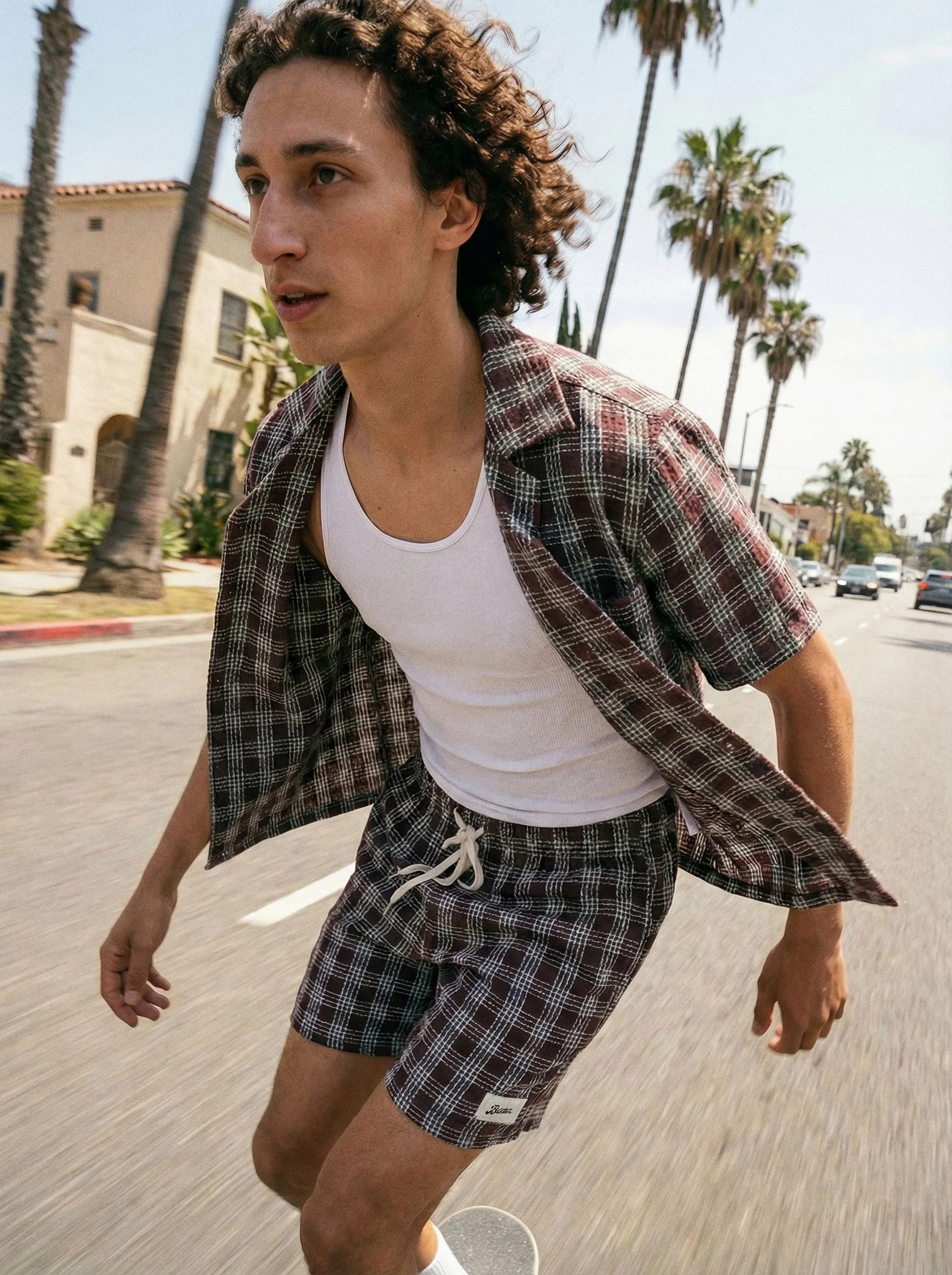 A young man with curly hair skateboarding on a street lined with palm trees, wearing a white tank top, plaid shirt, and plaid shorts.  AI fashion editorial image.Polo Ralph Laurent.