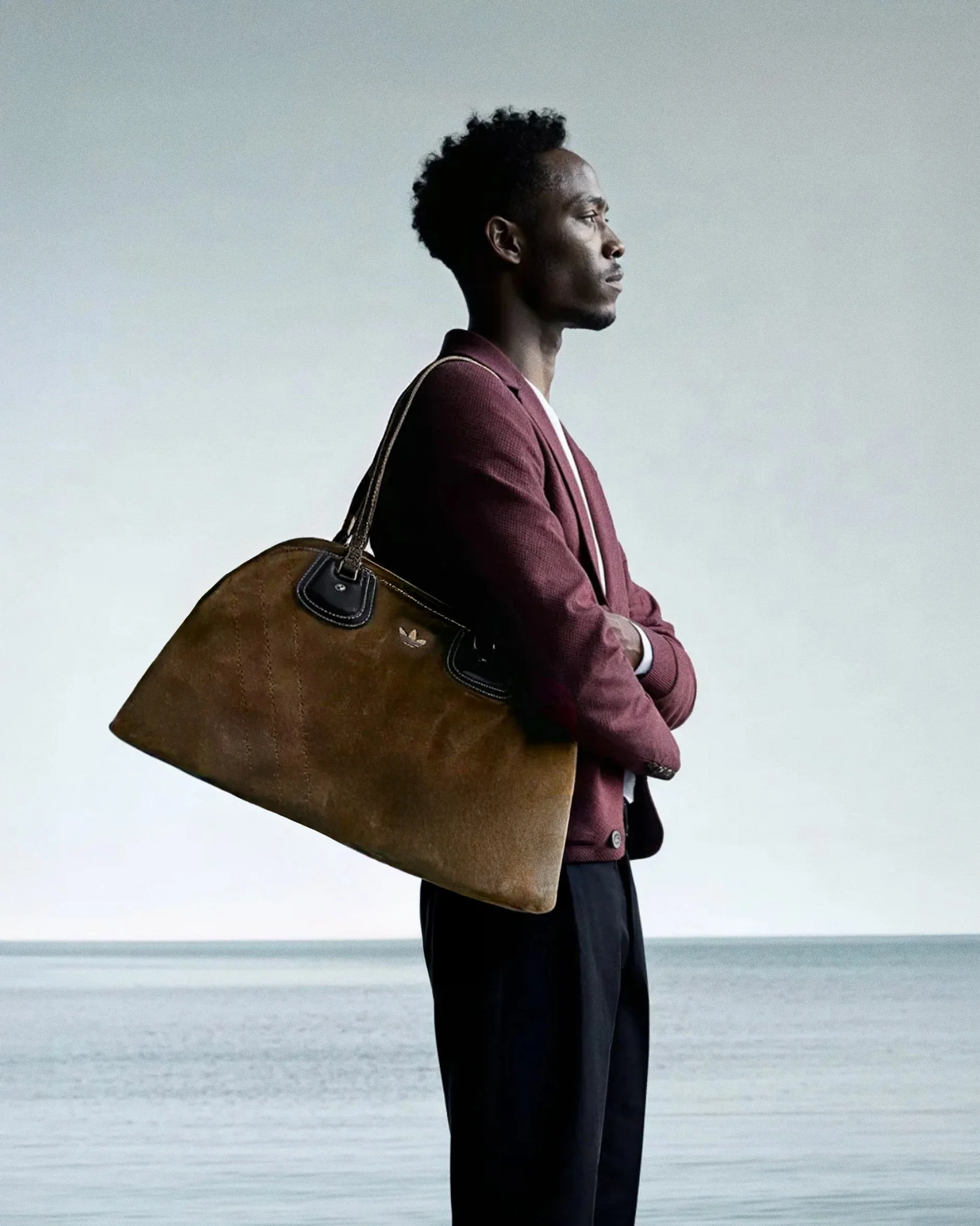 A man with dark skin and curly hair stands by a body of water, wearing a maroon blazer and black pants, carrying a large brown bag over his shoulder, looking to the side.