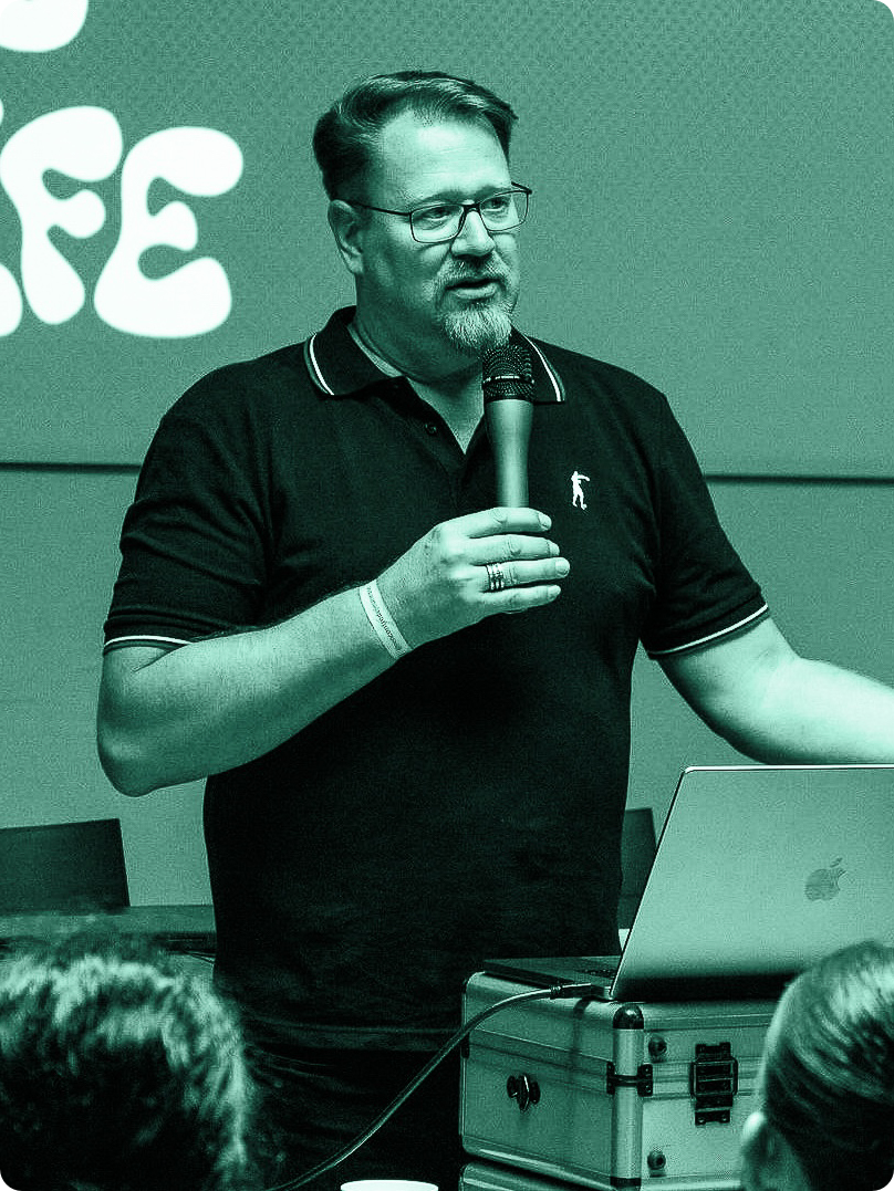 Ein Mann spricht vor einer Gruppe, hält ein Mikrofon in der Hand und steht neben einem geöffneten Laptop, während andere Personen im Vordergrund zuhören.