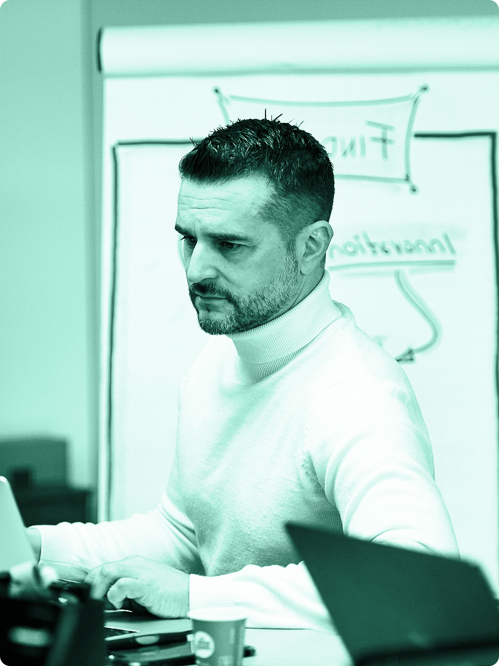 Mann arbeitet an einem Laptop in einem Besprechungsraum, im Hintergrund ein Whiteboard mit 'Finish' und 'Interpretation' geschrieben.