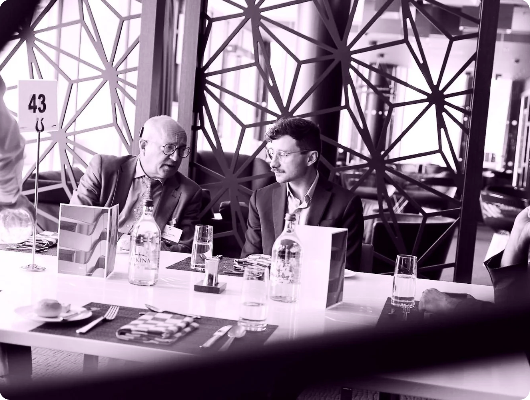 Drei Männer in Anzügen sitzen an einem Tisch in einem Restaurant oder Konferenzraum, diskutieren miteinander. Der Tisch hat Flaschen Wasser, Gläser, Menükarten und Besteck. Im Hintergrund sind dekorative Gitterwände. Der Raum wirkt modern und hell.