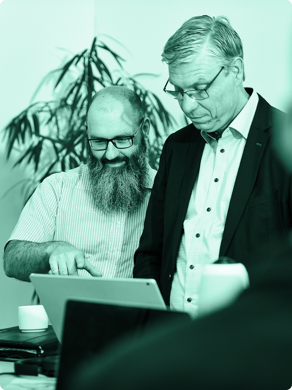 Zwei Männer in einem Büro, die auf einen Laptop schauen und diskutieren, während im Hintergrund eine Pflanze steht.