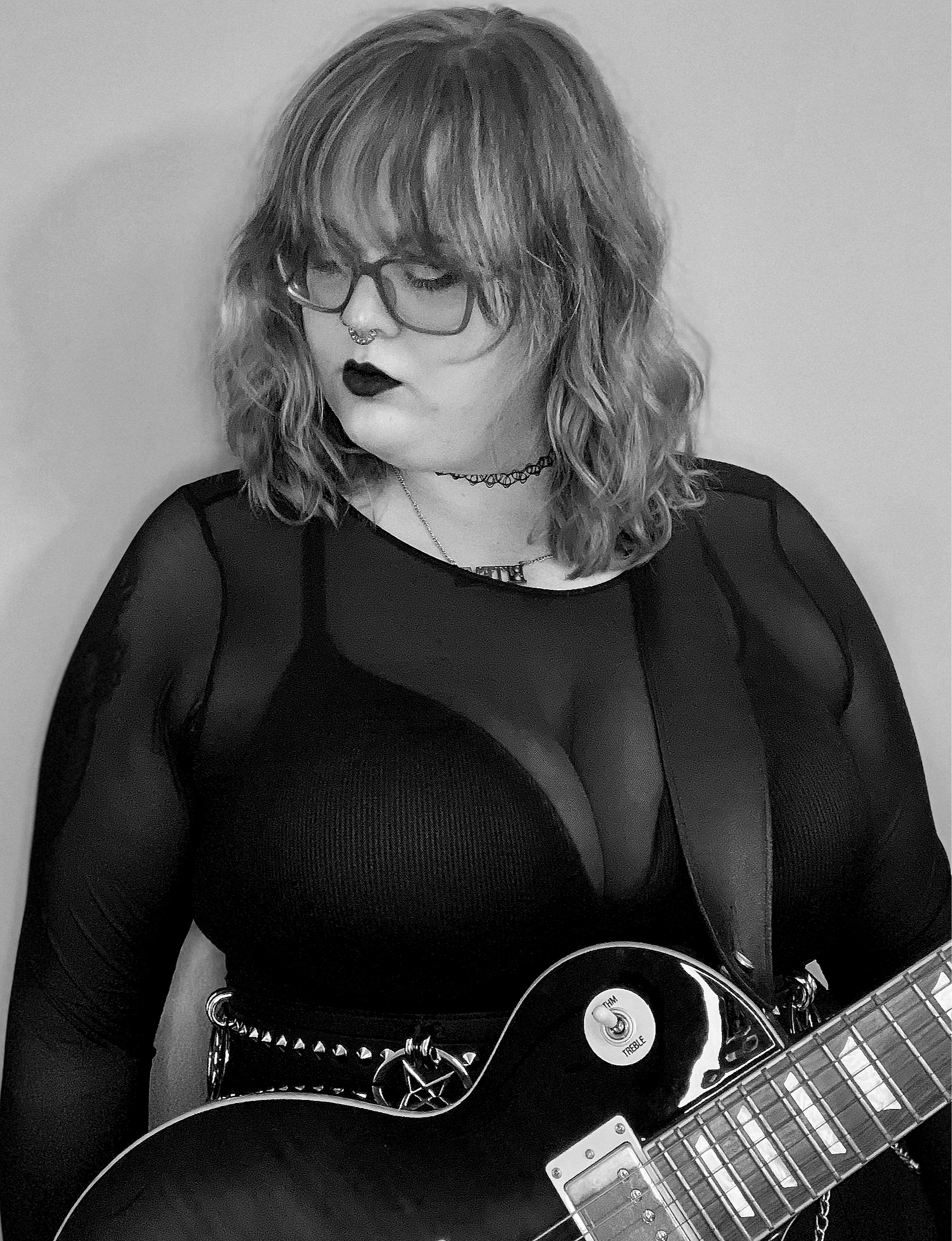 Black and white photo of a feminine person with wavy hair and a mesh long sleeved shirt looking down and to the side whilst holding a Les Paul guitar