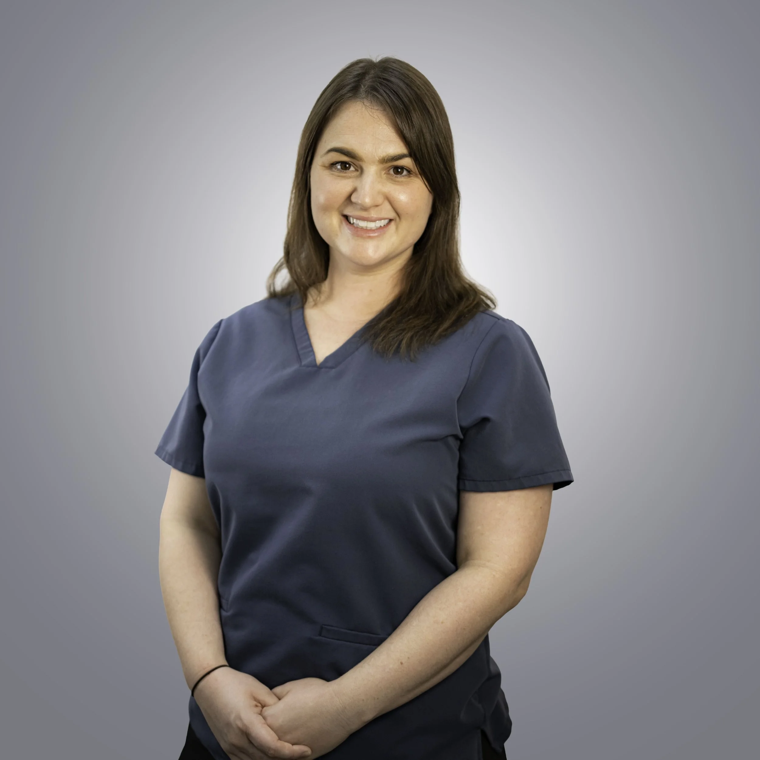 A woman with brown hair wearing navy blue scrubs, smiling with folded hands, standing against a light gray background.