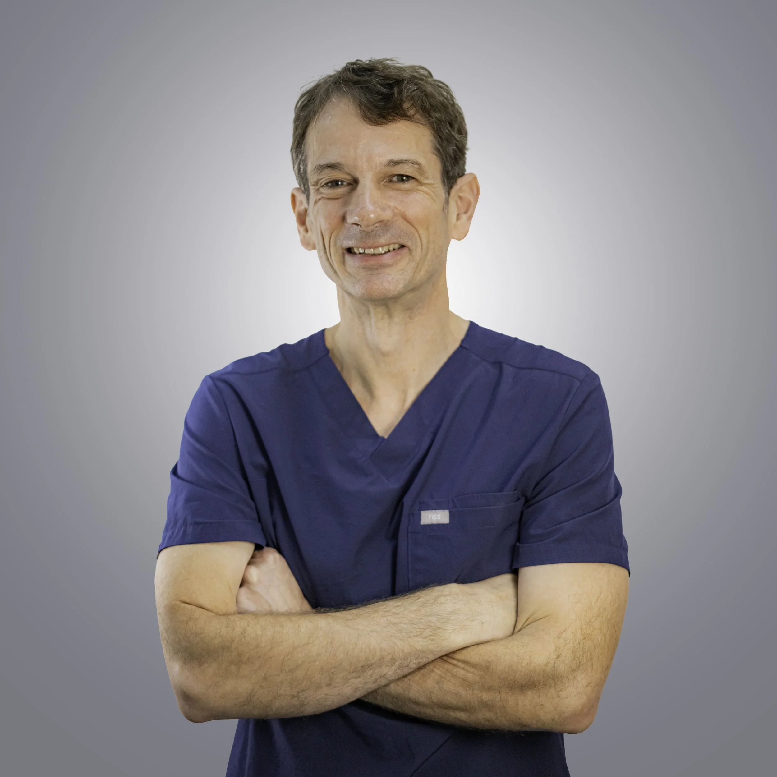 A smiling male doctor or medical professional in blue scrubs, with arms crossed and a friendly expression, standing against a gray background.