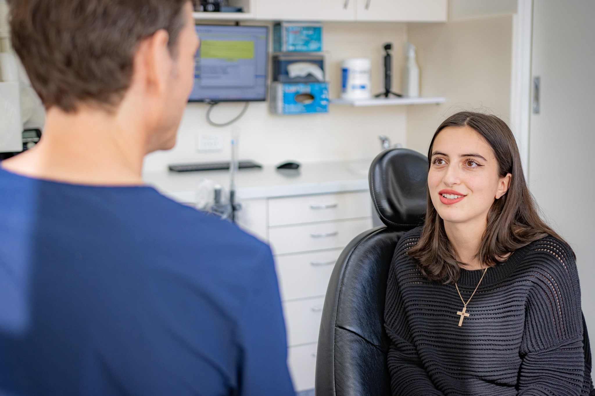 A young woman with long brown hair and a cross necklace smiling and talking to a man at a medical or dental office, with medical equipment in the background.