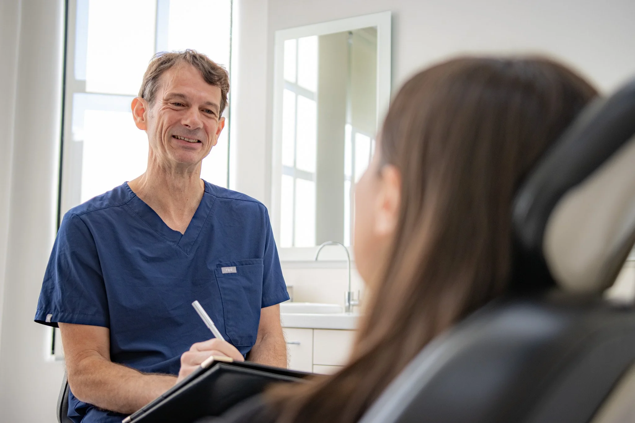 A male doctor in blue scrubs talking to a female patient sitting in a medical chair.