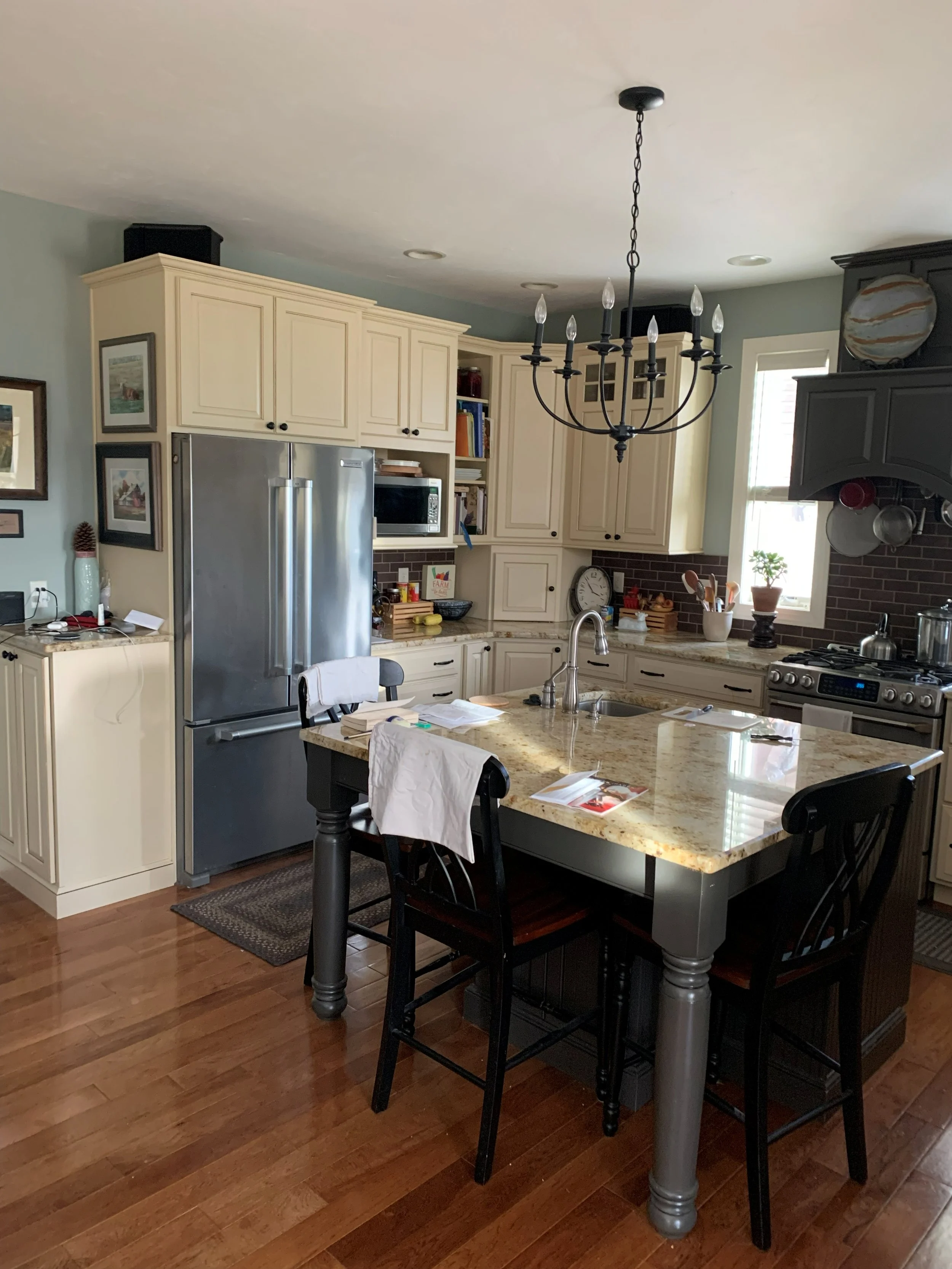 R.E. Arnold's Custom Construction. Kitchen install with cream-colored cabinets, a stainless steel refrigerator, a kitchen island with a beige marble countertop, and a black chandelier hanging from the ceiling.