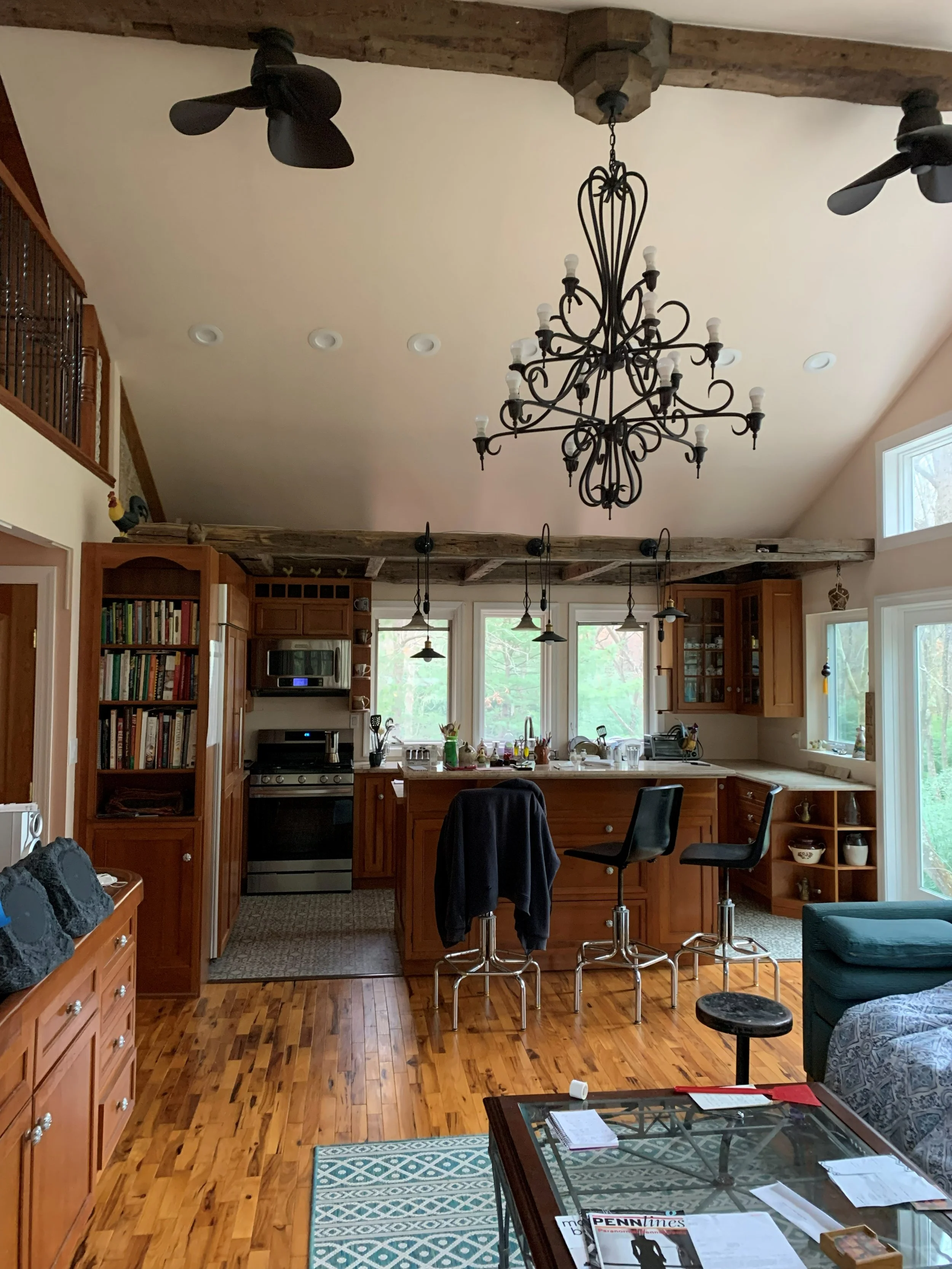 R.E. Arnold's Custom Construction. A cozy kitchen with wooden cabinets and a large window behind the sink, featuring a rustic wood beam ceiling and a black chandelier. 