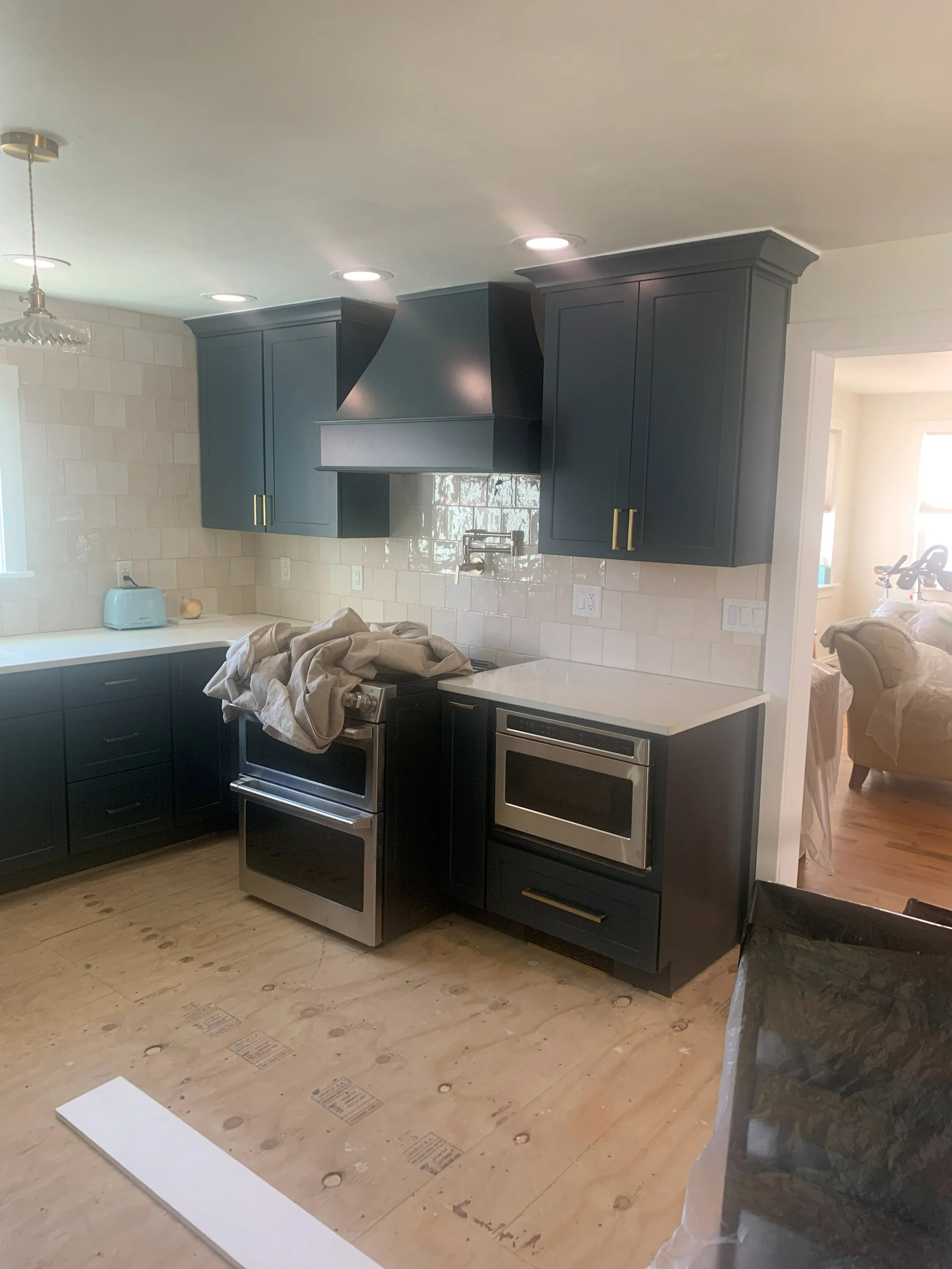 R.E. Arnold's Custom Construction. Modern kitchen with dark navy cabinets, stainless steel oven, and white countertops. The floor is unfinished, with plywood visible, and there is a cloth draped over the oven.