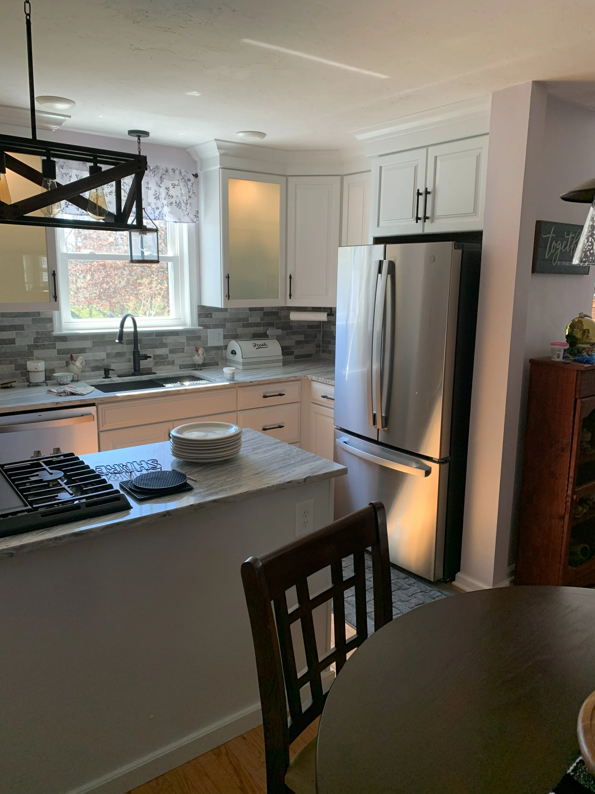 Kitchen with white cabinets, stainless steel refrigerator, gray tile backsplash, window above sink, and a marble island with stacked plates.
