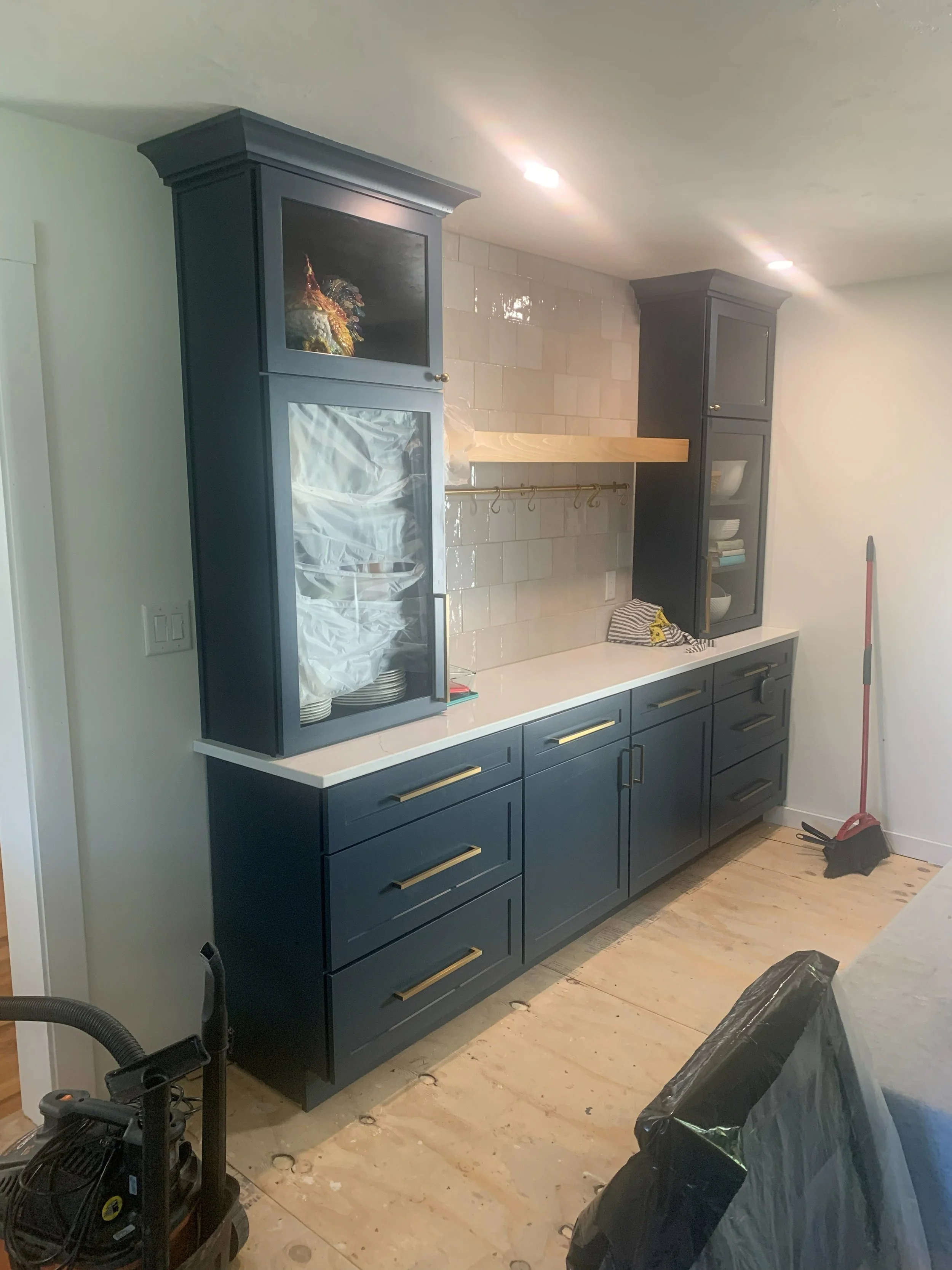 R.E. Arnold's Custom Construction. Modern kitchen cabinet with blue paint, gold handles, white countertop, open shelf, and light-colored tiled wall, with cleaning tools and plastic-covered furniture in the room.