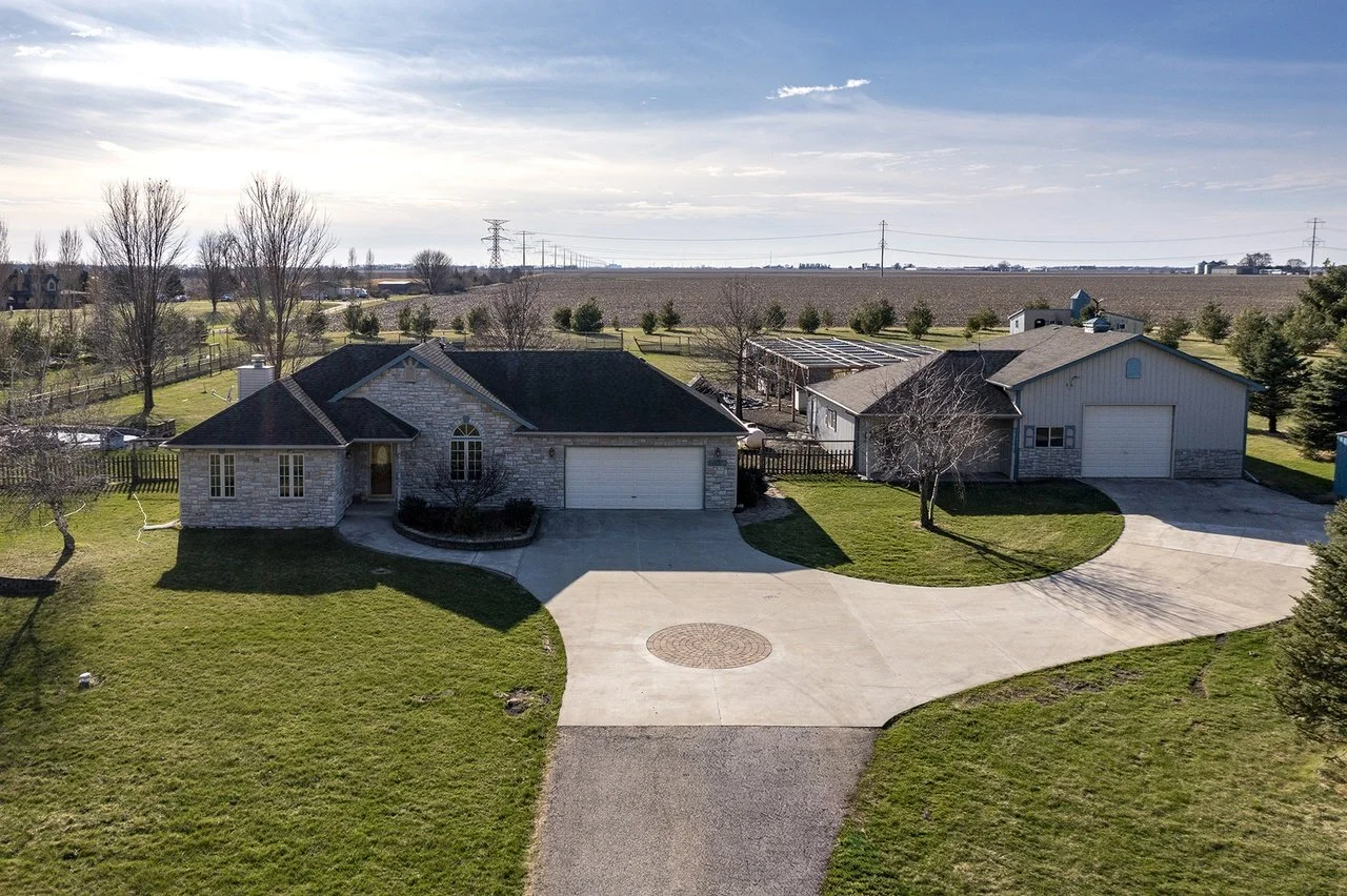 An aerial view of a ranch-style house in Verona, IL, with a large front yard, garage, and pole building. Surrounding the house are trees, a fence, and open farmland in the background.