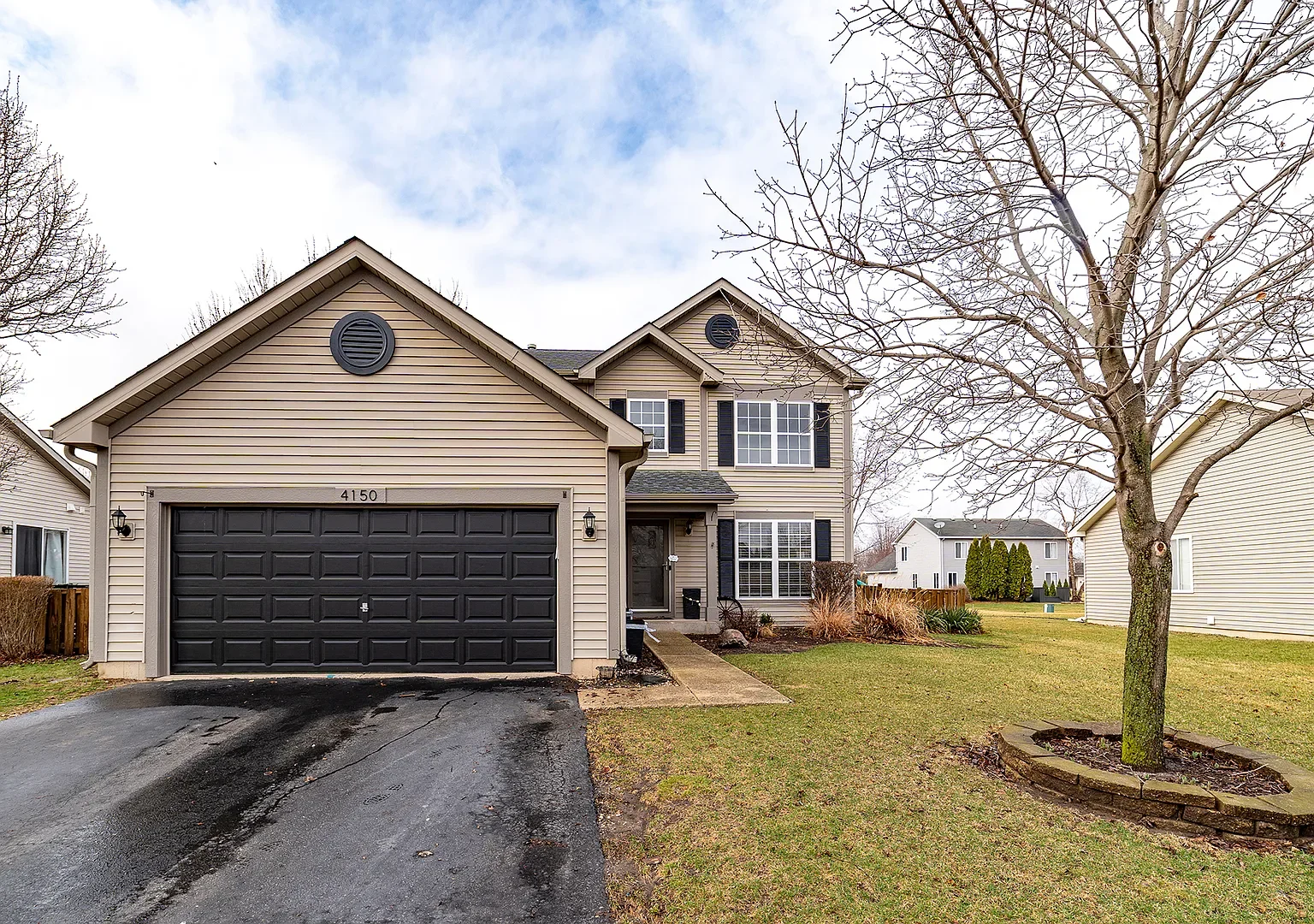 Two-story house with beige vinyl siding, shutters, and a garage door leading to the driveway. There is a large tree in the front yard surrounded by a raised circular flower bed. Other houses are visible in the background.