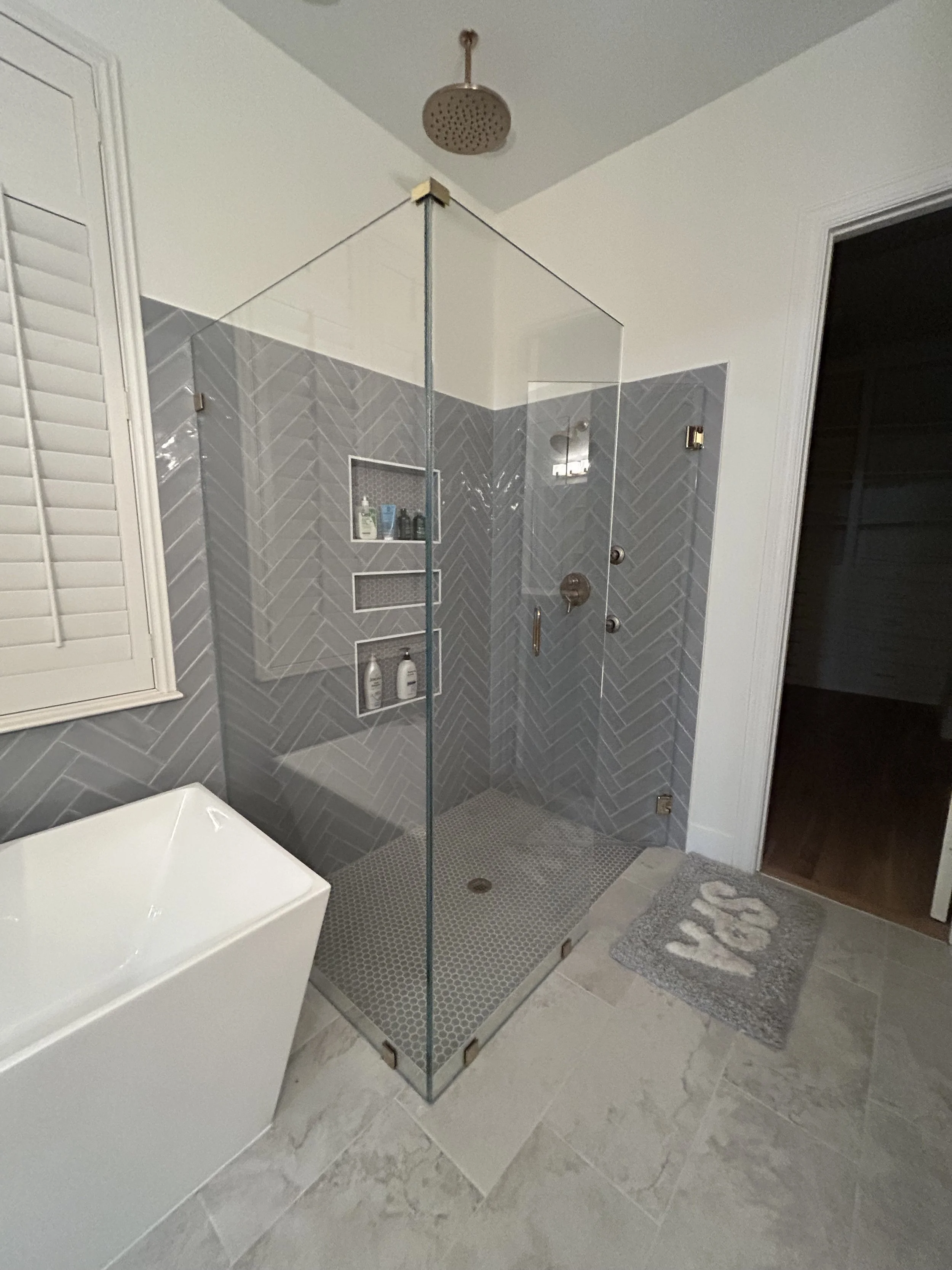 Custom Shower Tile Design – Prosper, TX. Clean, modern bathroom design featuring herringbone wall tile, mosaic shower floor, and frameless glass enclosure for a bright, elegant new-build space.