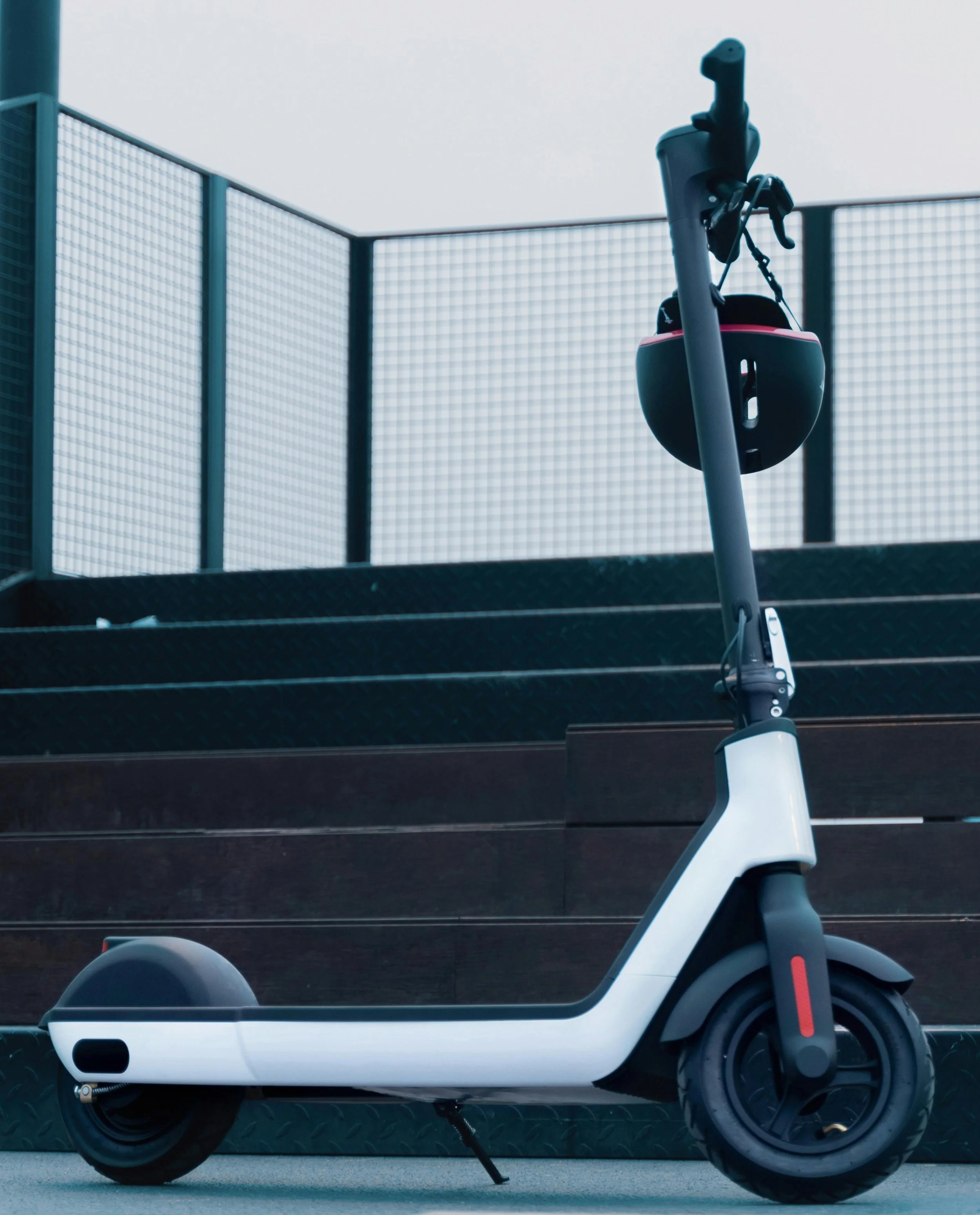Electric scooter parked near stairs with a black riding helmet hanging on the handlebar, outdoors near a metal fence.