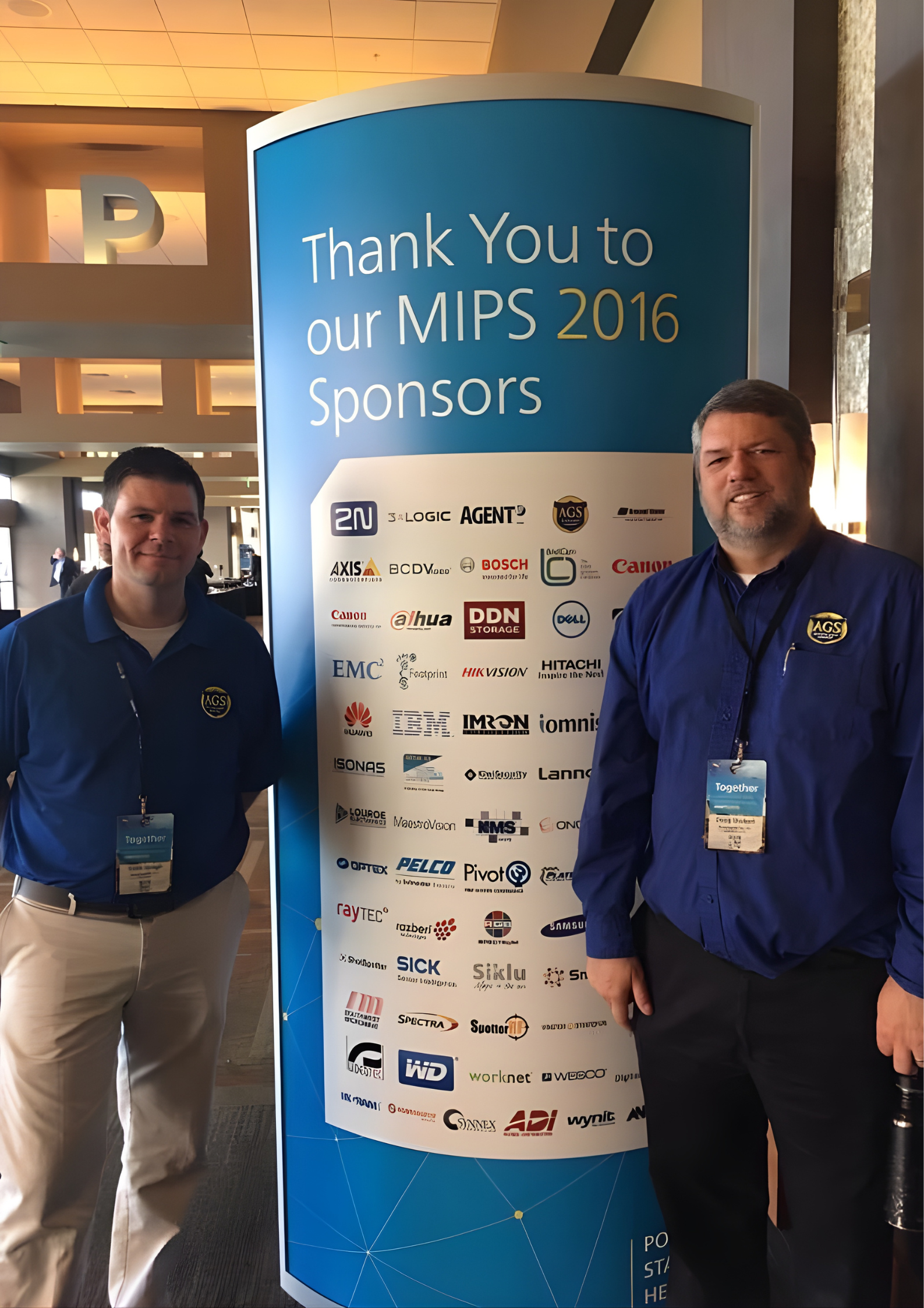 Two men standing next to a large blue sign with logos of sponsors for MIPS 2016, a conference or event, inside a building with a modern interior.