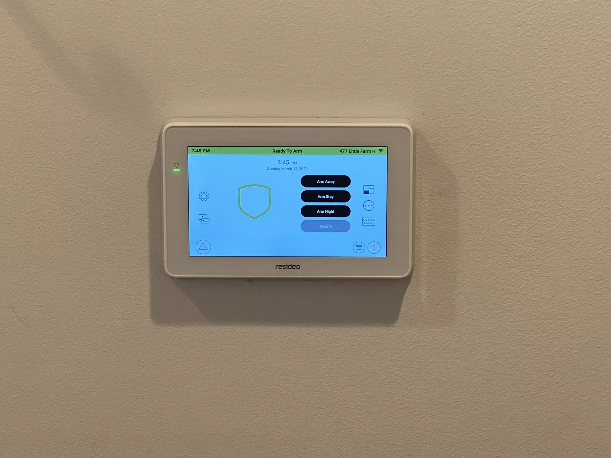 A home security touchscreen panel mounted on a beige wall, displaying options to arm the security system in away, stay, or night modes.