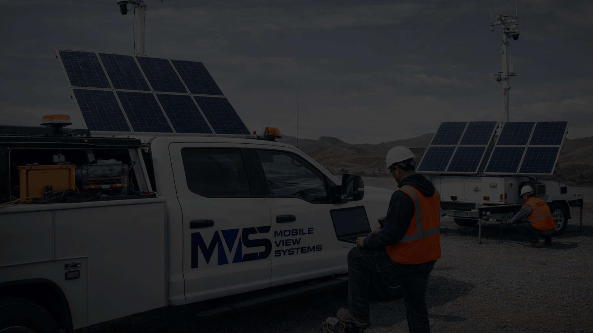 Technicians working outdoors near a white vehicle with solar panels installed on it, in a desert landscape with mountains in the background.