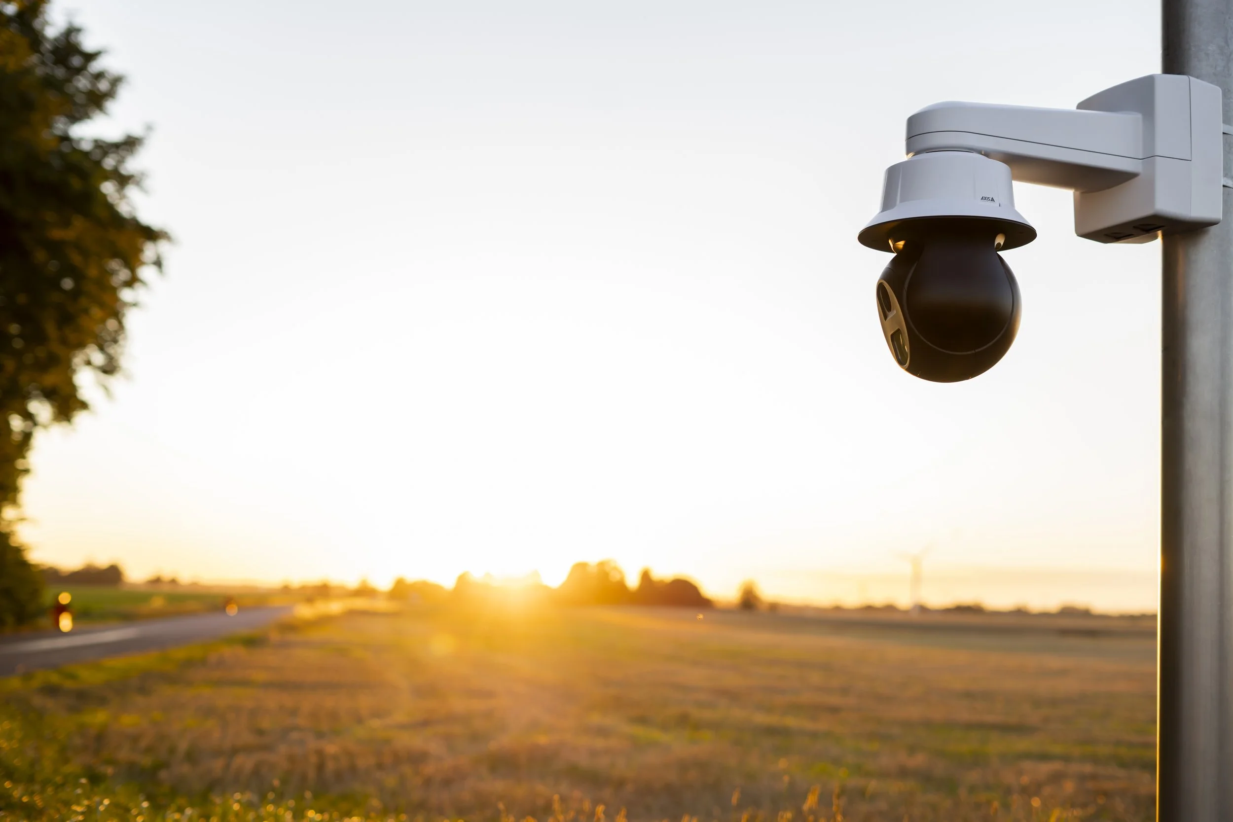 A surveillance security camera mounted on a pole, with a sunset or sunrise in the background over a rural landscape with fields and trees.