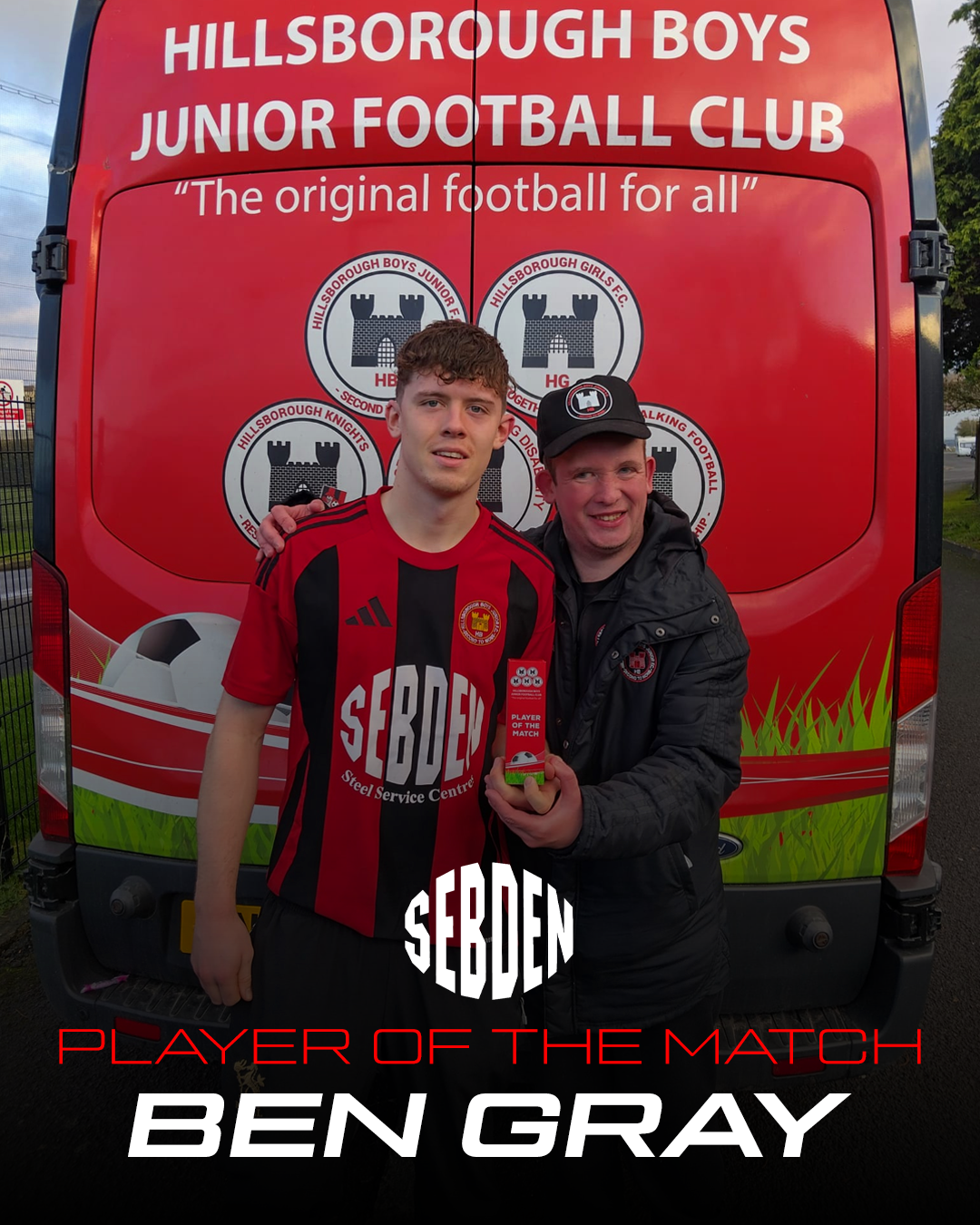 A young soccer player in a red and black jersey standing next to a man in a black jacket and cap, holding a small trophy. They are standing in front of a red vehicle with 'Hillsborough Boys Junior Football Club' logos and text, celebrating Ben Gray as Player of the Match.