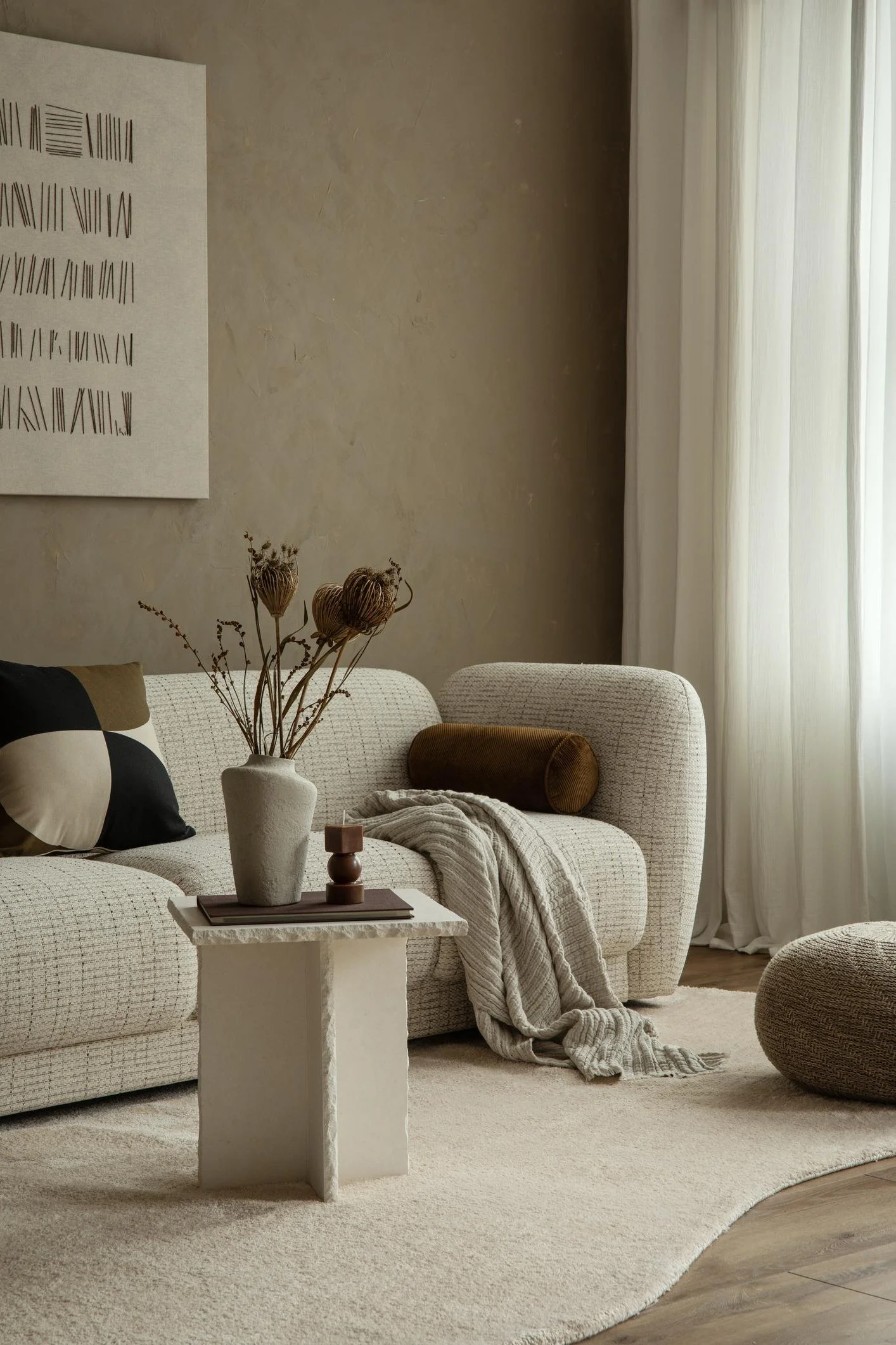Living room with beige textured sofa, a brown bolster pillow, a gray throw blanket, and a small white stone coffee table with a vase of dried flowers and geometric decor. Light-colored curtains hang in the background.