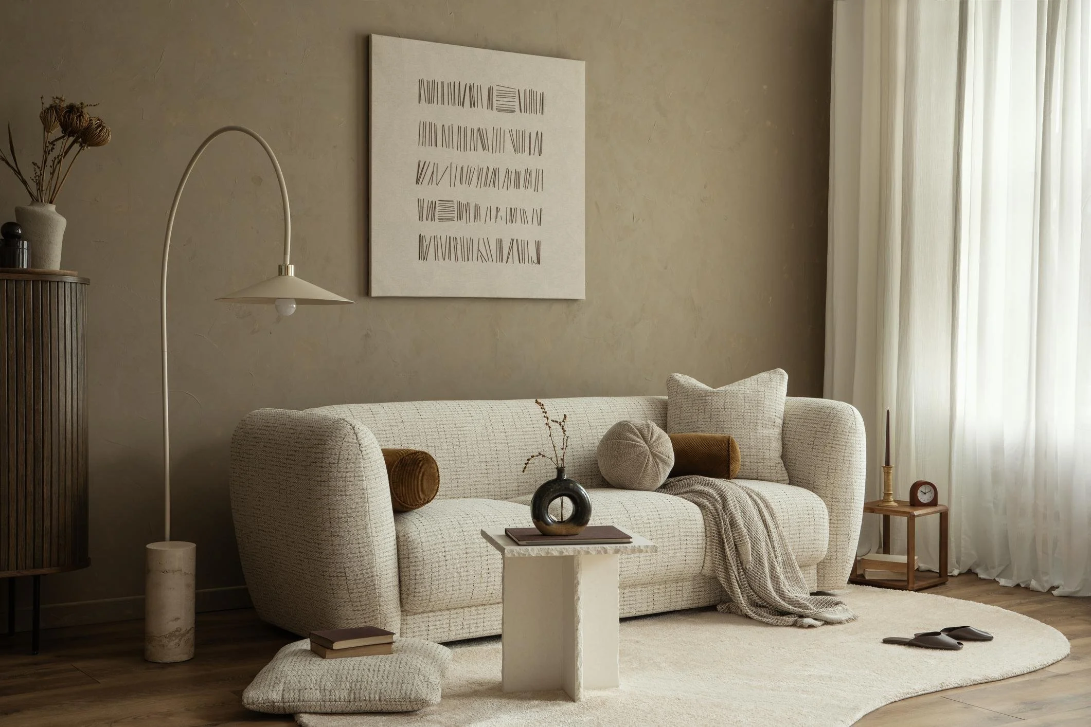 Living room with a light-colored sofa, a white coffee table, and a beige rug. Decor includes a black vase with dried flowers, books, a small table with a clock, and a tall side table with a lamp and curtain. Wall art with abstract lines hangs above the sofa.