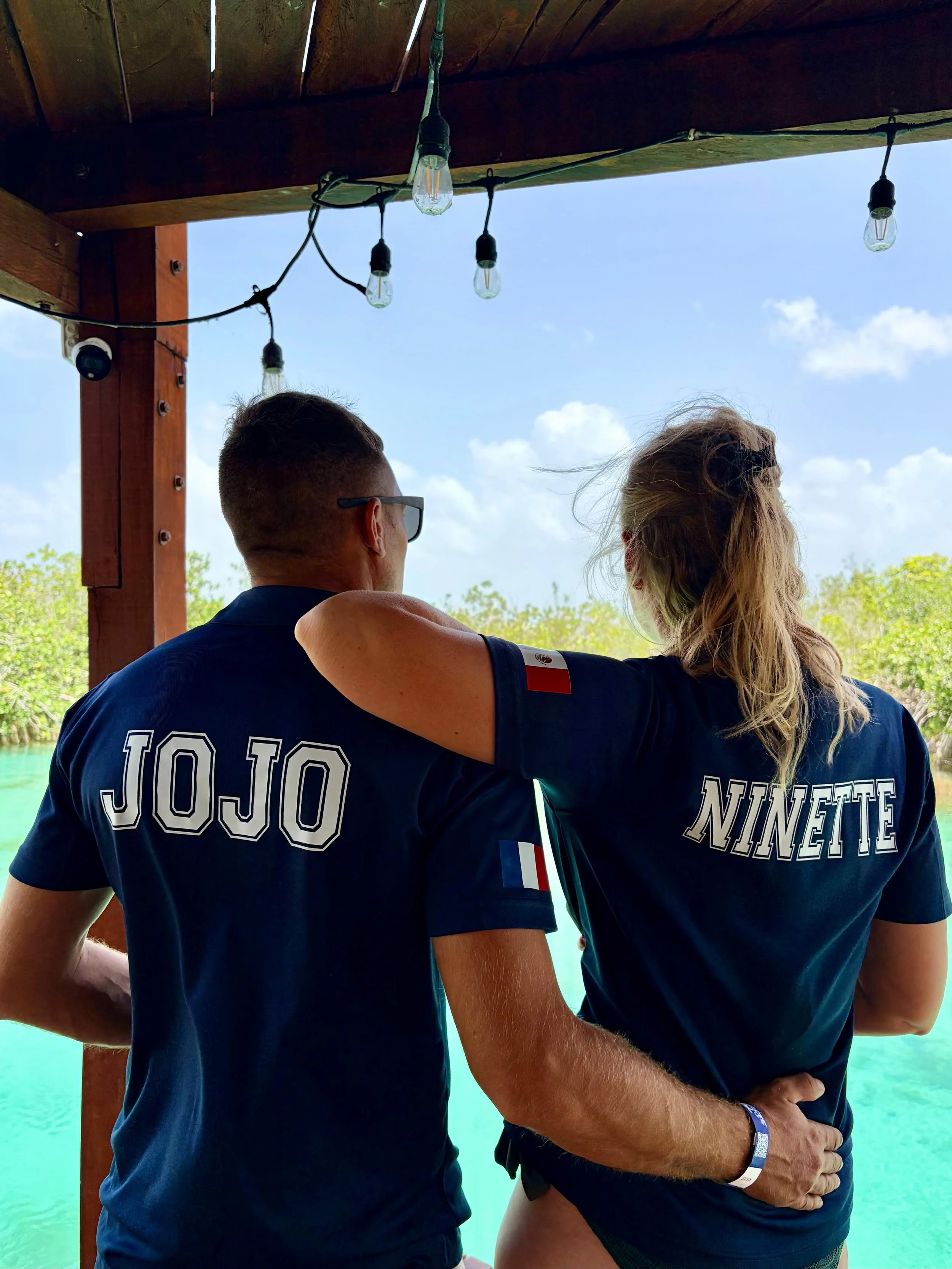 Deux personnes portent des T-shirts bleus avec les inscriptions 'JOJO' et 'NINETE' dans un environnement extérieur avec ciel bleu et eau turquoise derrière elles.