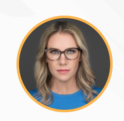 A woman with blonde wavy hair, wearing glasses, and a blue top, looking directly at the camera against a dark background.