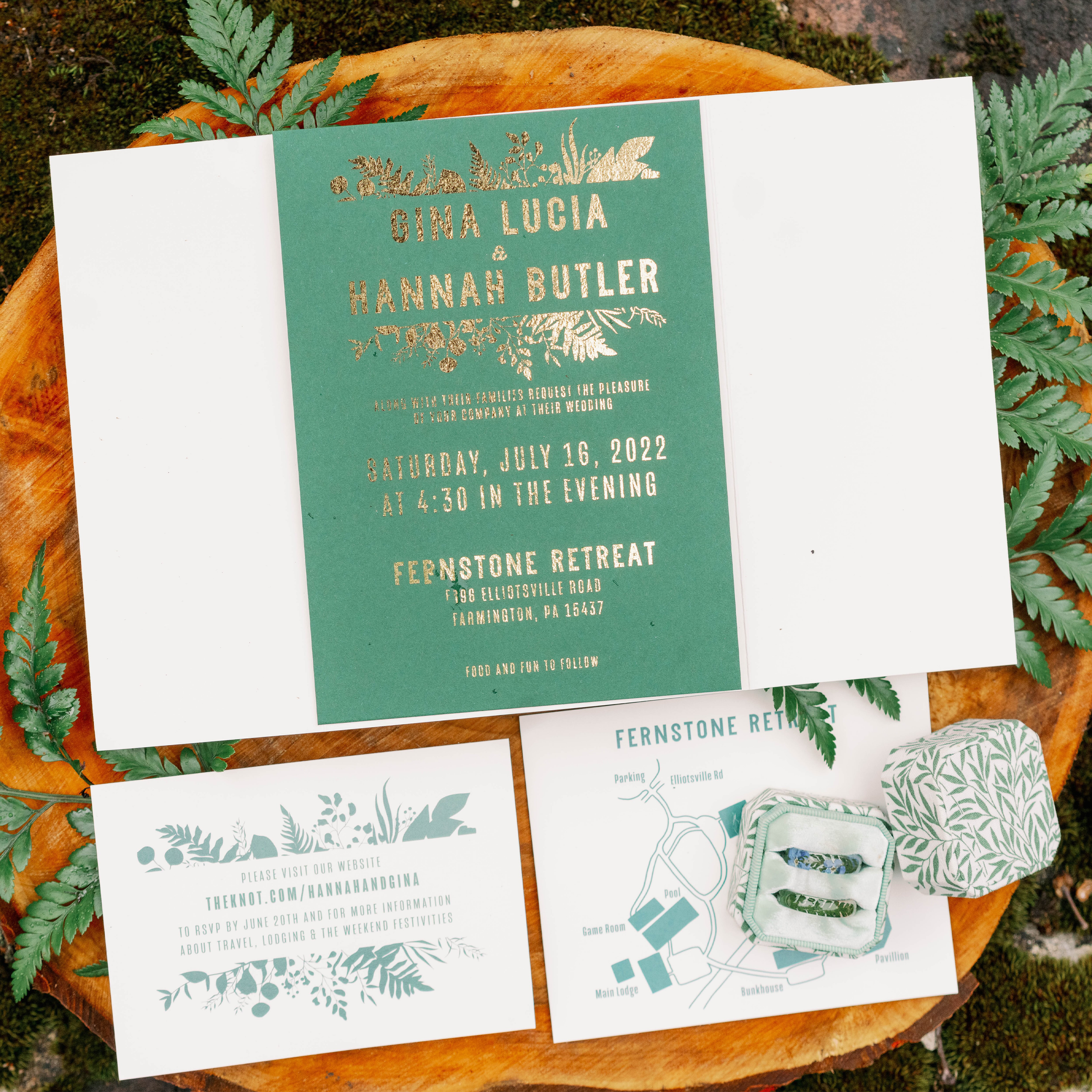 Wedding invitation suite with green and white cards, including a gold-foiled invite, a map, and RSVP details, arranged on a wooden slice with fern decorations and a small wrapped gift.