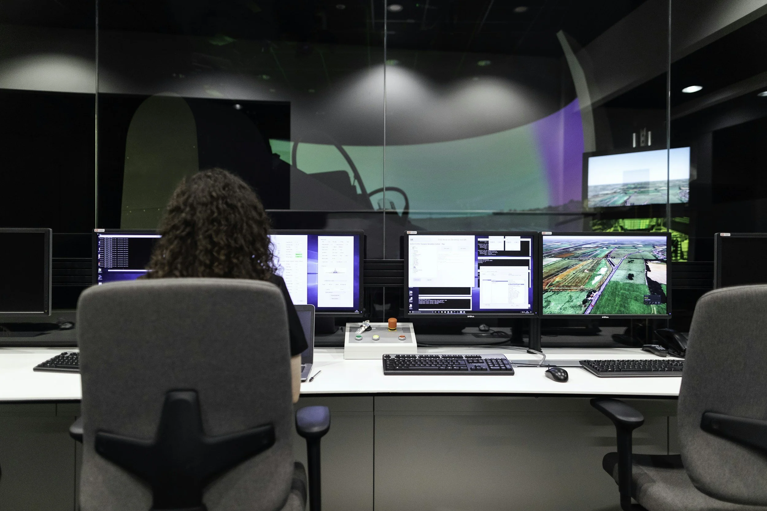 Control room with multiple computer monitors displaying maps and data, a person working at a desk with a control panel, inside a high-tech facility with a large glass wall showing a visual display.