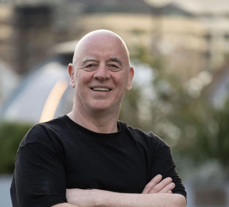 A smiling bald man wearing a black shirt with arms crossed, standing outdoors with blurry background of trees and buildings.