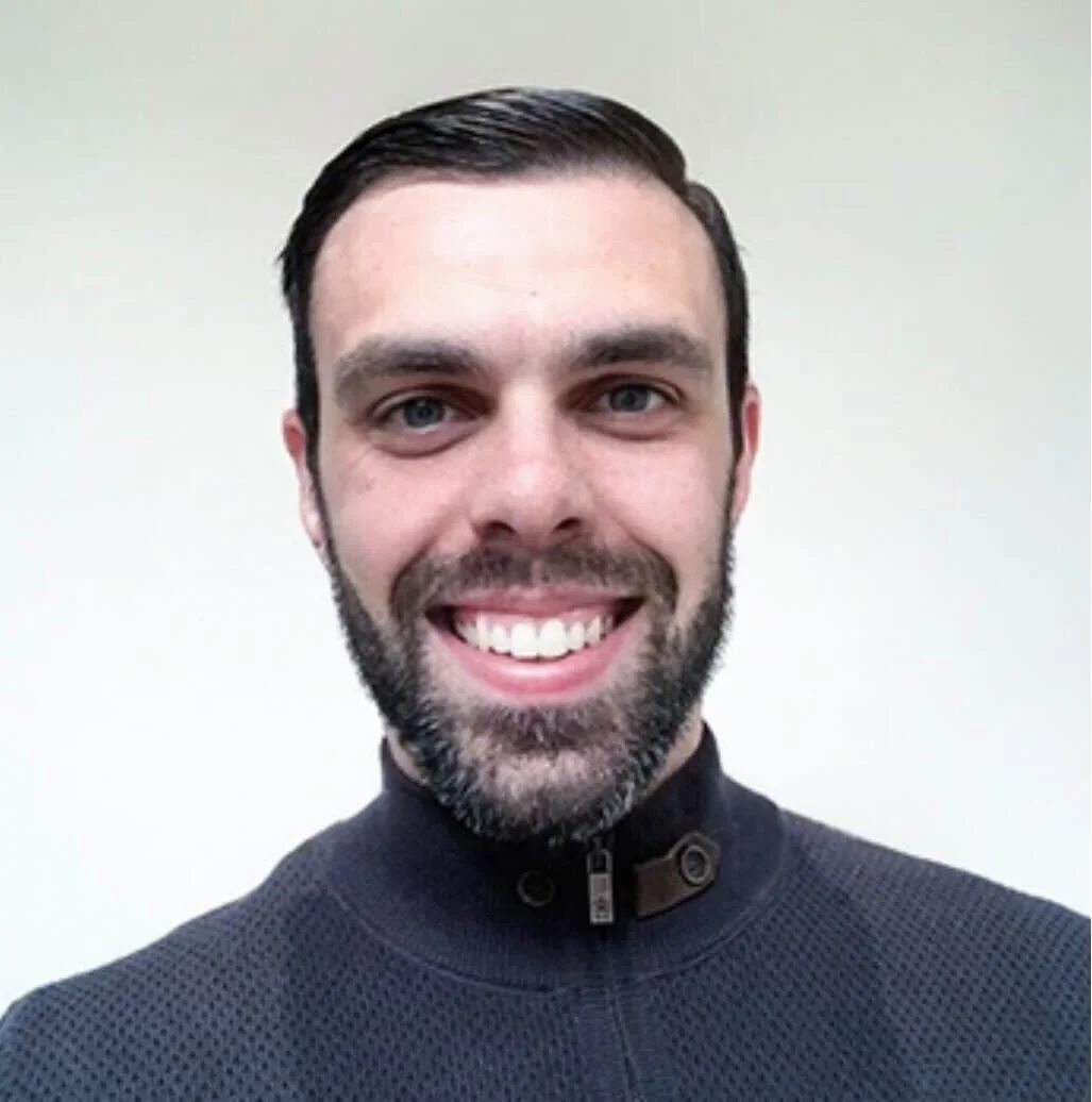 A smiling man with dark hair, beard, and blue eyes, wearing a dark zip-up jacket, standing against a plain off-white background.
