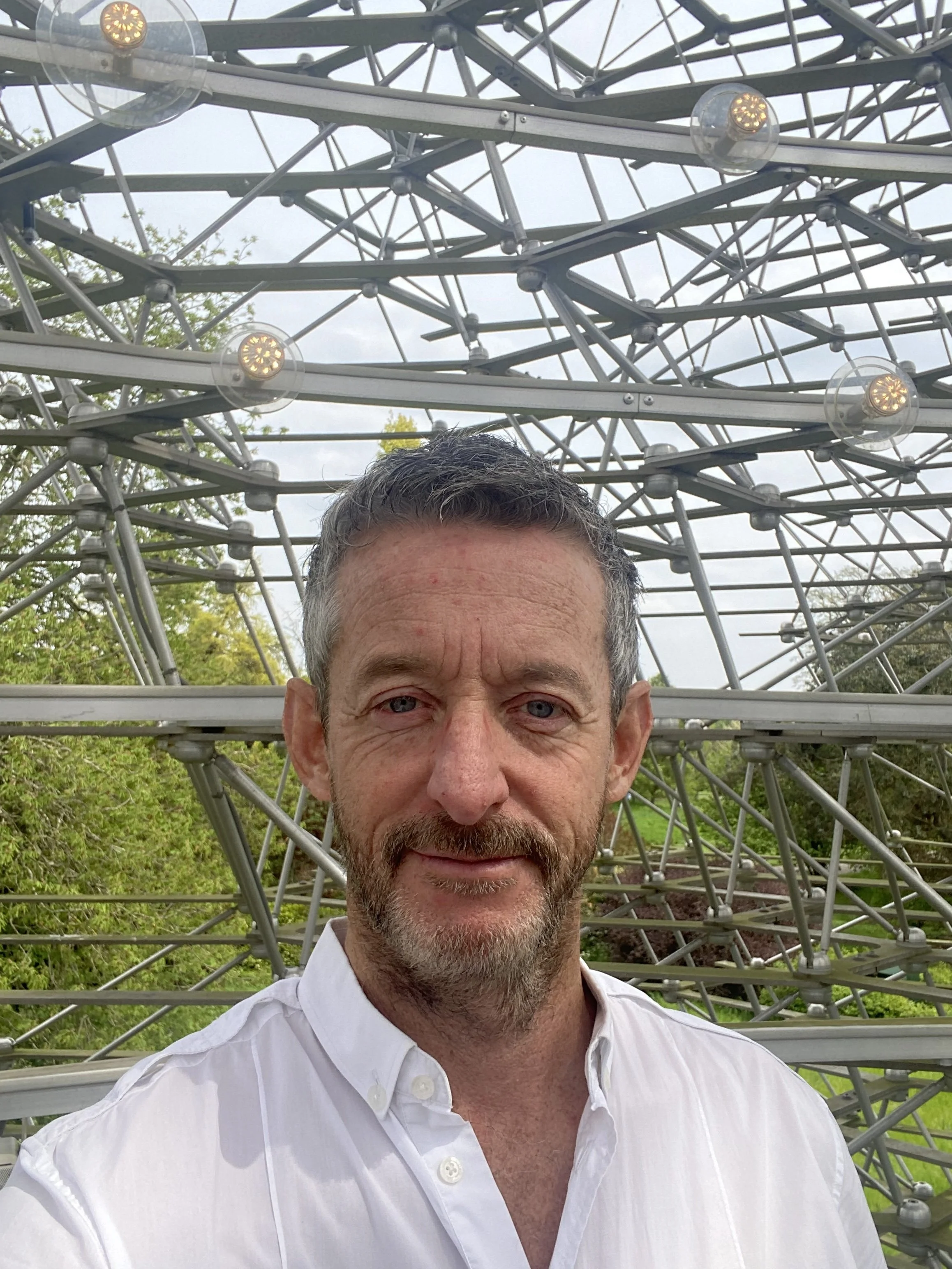 A man with short, dark hair and a beard smiling in front of a geodesic dome structure outdoors on a cloudy day.