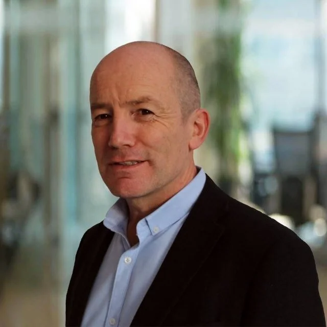 A middle-aged man with a shaved head wearing a light blue dress shirt and black blazer, standing indoors with a blurred background of windows and plants.