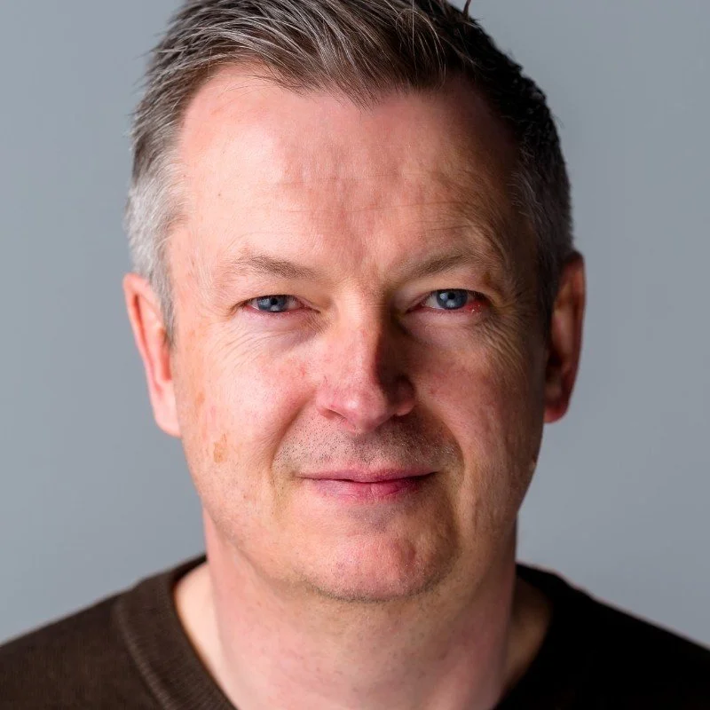 A middle-aged man with short brown hair and blue eyes, wearing a dark sweater, smiling slightly against a plain gray background.